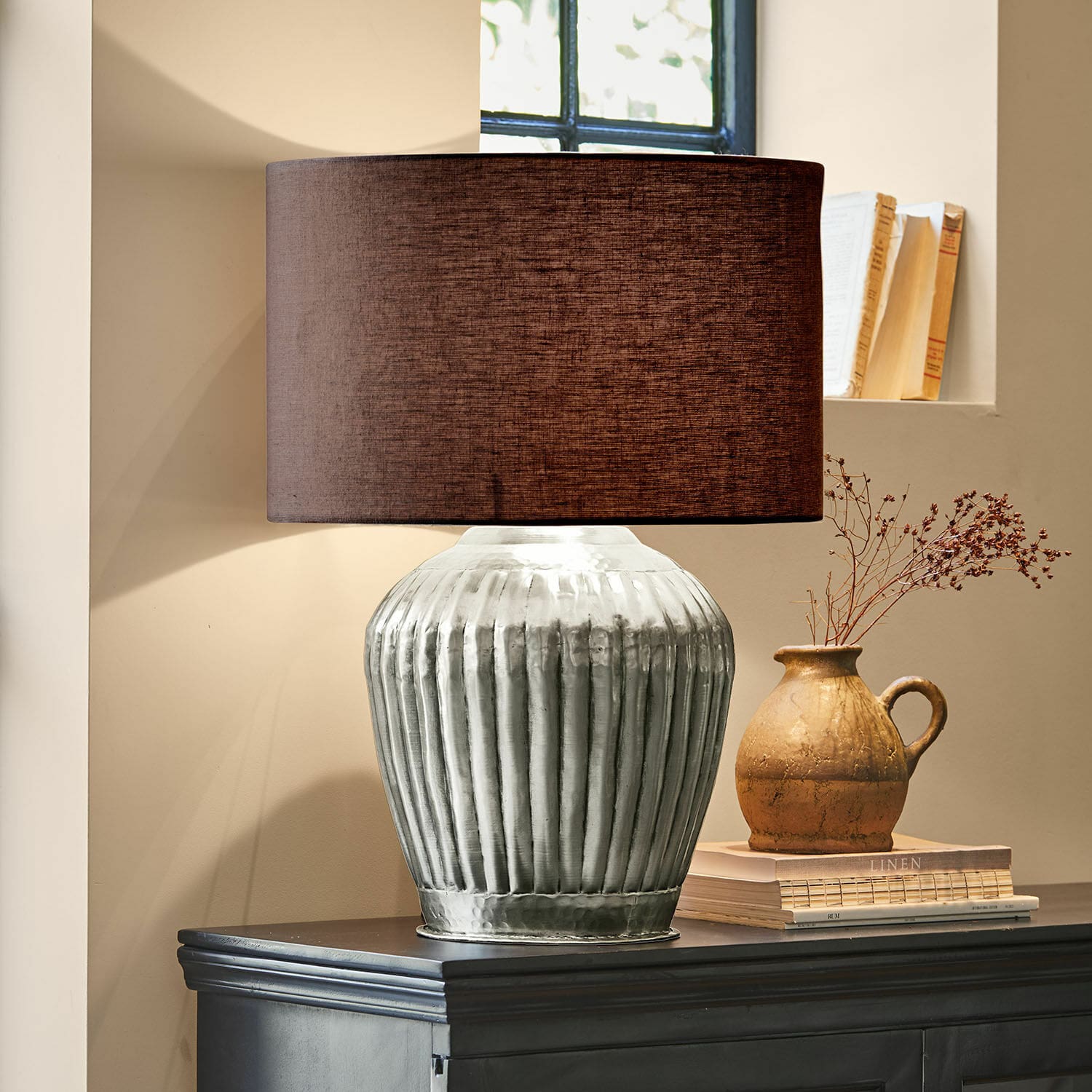 Stolní lampa Fauville Stolní lampa se stříbrnou kovovou základnou a hnědým stínidlem na tmavé komodě