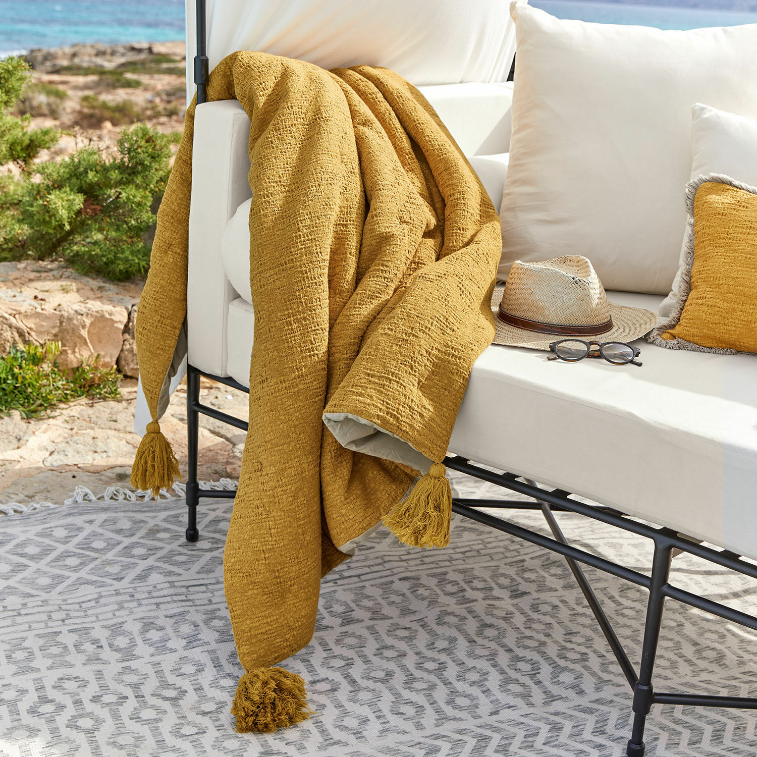 Pléd Rabea Žlutá deka s třásněmi na bílém venkovním gauči, dekorace terasy s brýlemi a kloboukem
