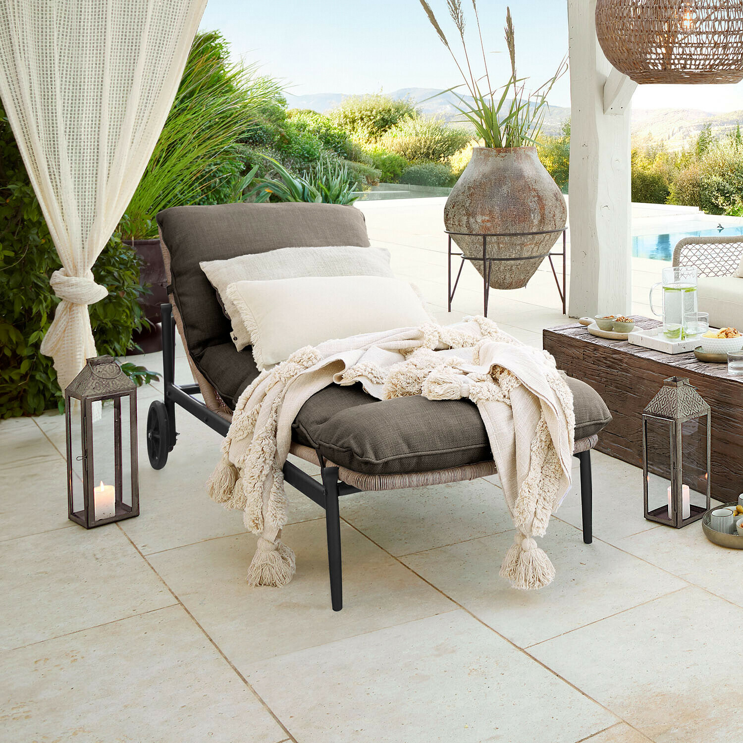 Zahradní lehátko s tmavě šedou látkou a krémovou dekou s třásněmi na terase s výhledem do zahrady