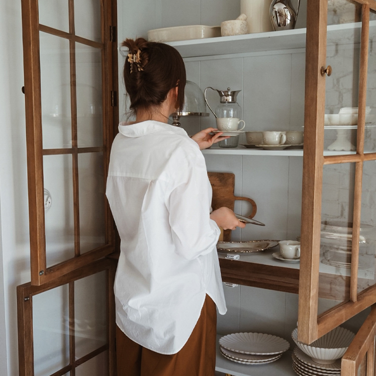 Žena v bílé košili bere porcelánový šálek ze dřevěné vitríny s nádobím
