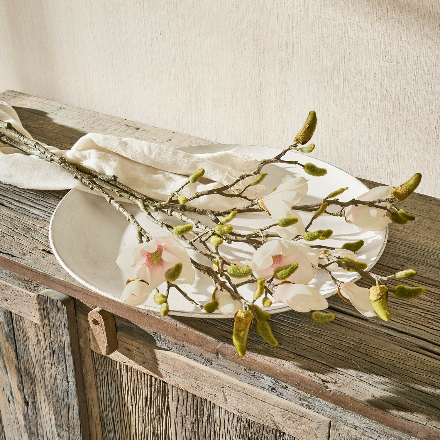 Větve magnólie s bílými květy a zelenými pupeny na rustikálním dřevěném stole s bílým talířem
