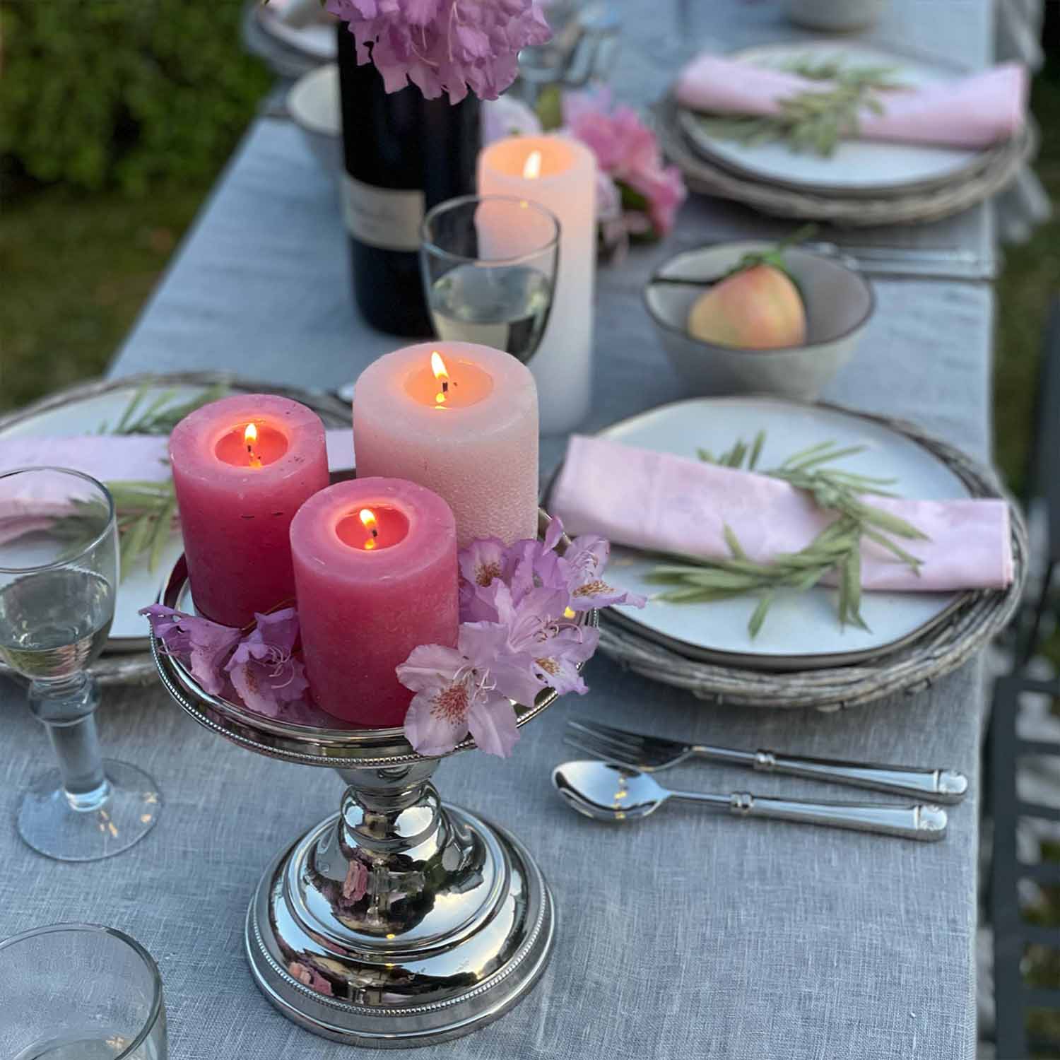 Stříbrný svícen s třemi hořícími růžovými a bílou svíčkou na šedém ubrusu s květinovou dekorací