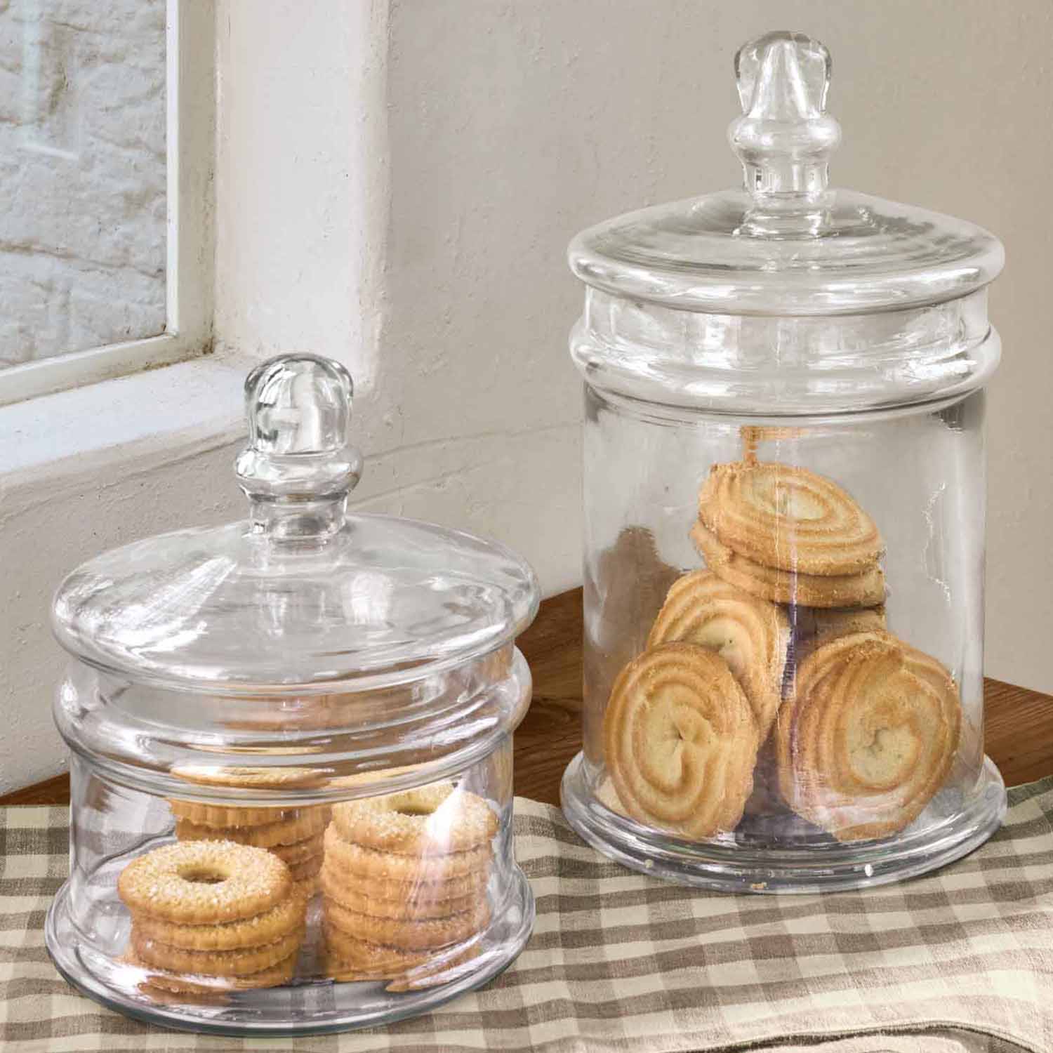 Dvě průhledné skleněné dózy s víčky plné sušenek na dřevěném stole s kostkovaným ubrusem