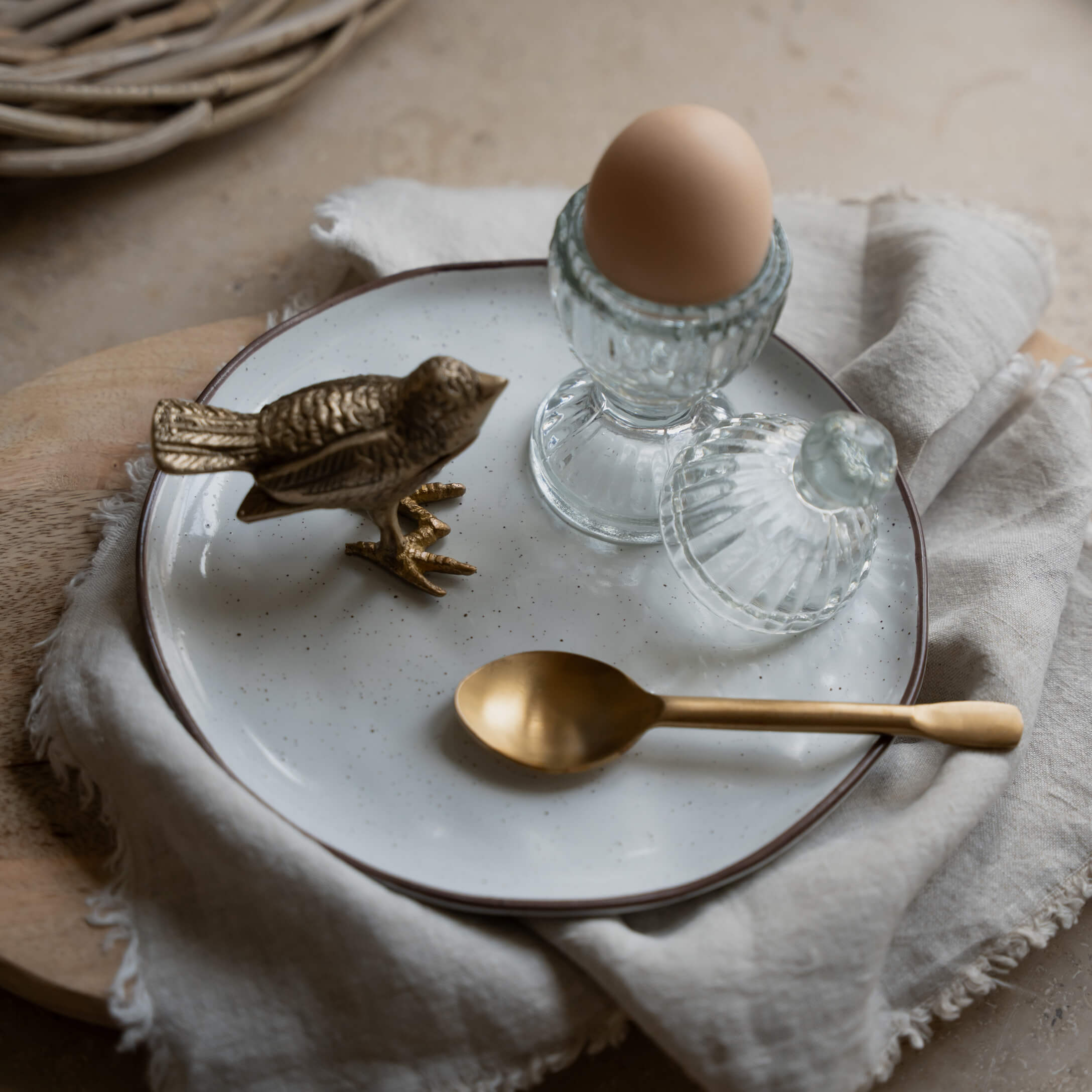 Skleněný stojánek na vejce s víčkem a hnědé vejce na bílém talíři s mosaznou lžičkou a soškou ptáka