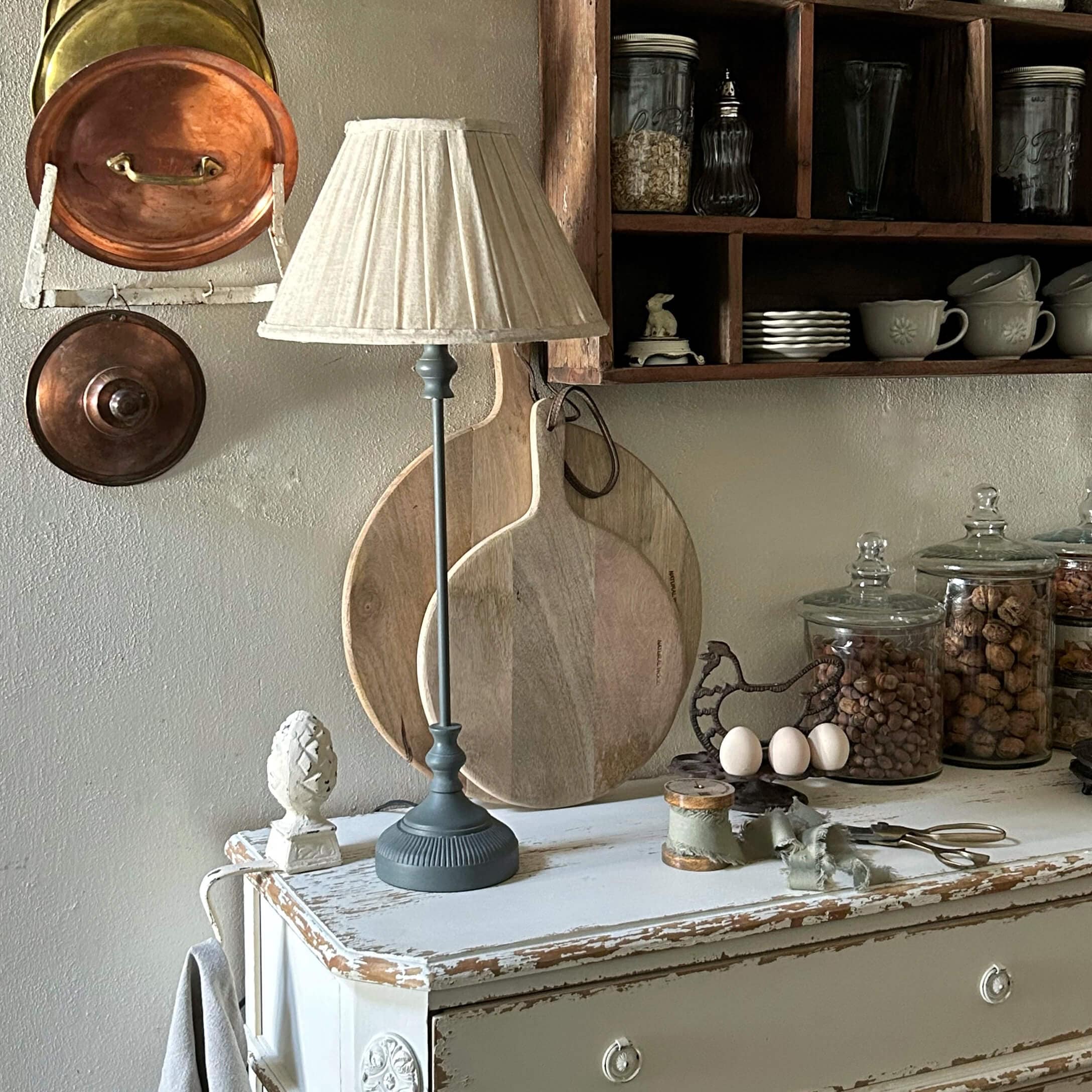 Stolní lampa se šedým podstavcem a krémovým stínidlem na bílém starobylém kredenci v rustikální kuchyni