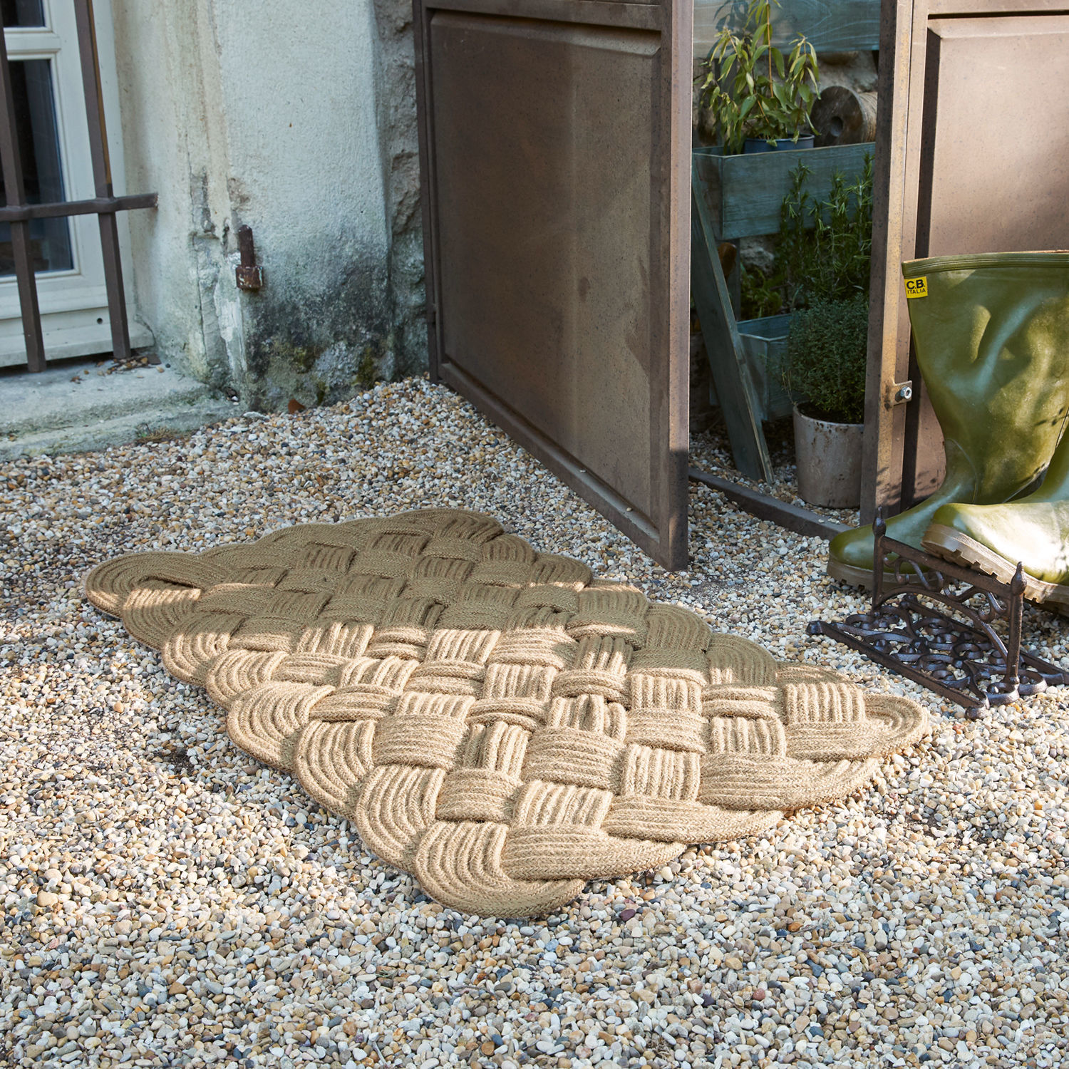 Béžová rohož z pleteného provazu na štěrkovém povrchu u otevřených dveří vedle zelených holínek
