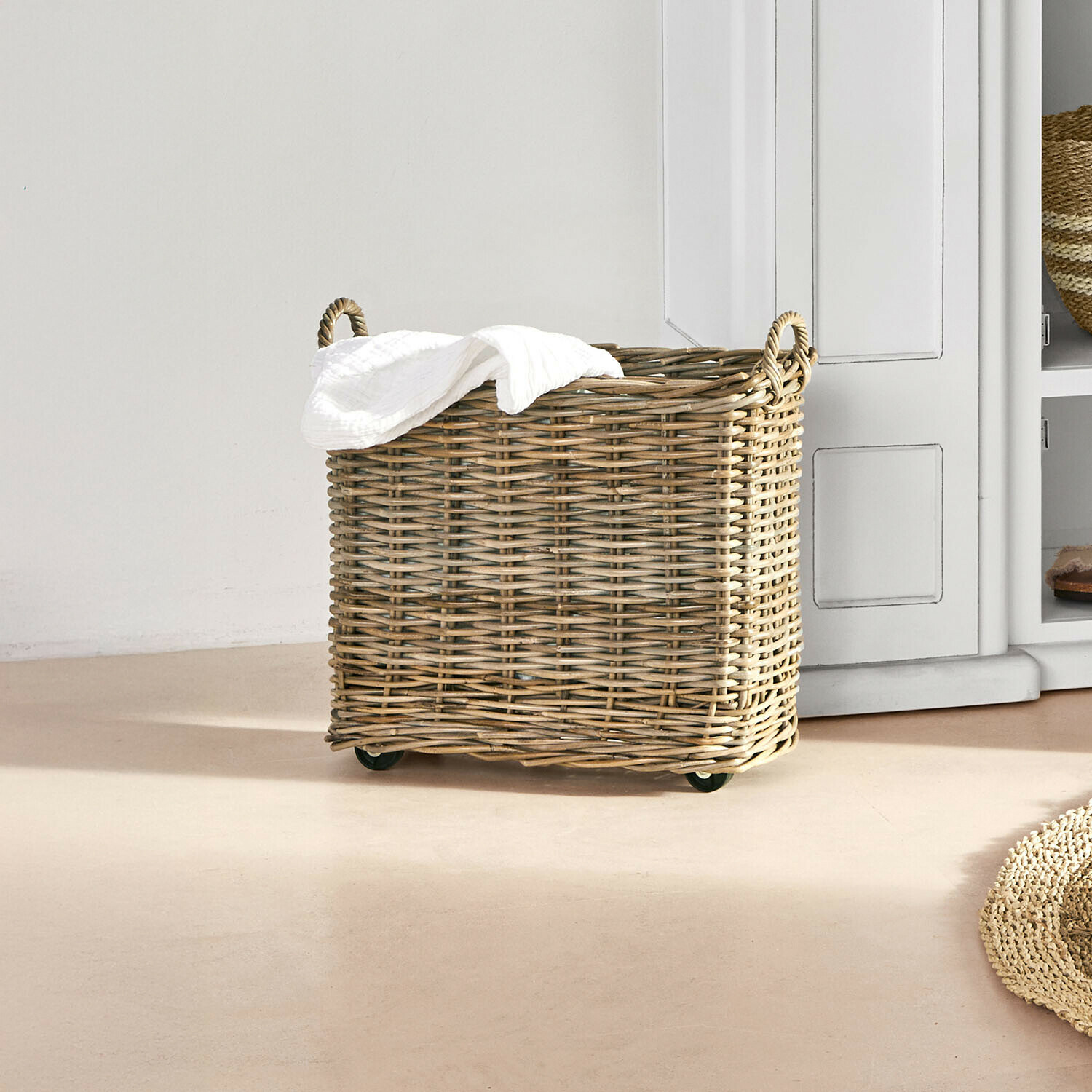 Proutěný koš na prádlo na kolečkách v přírodní barvě s bílým ručníkem v světlé místnosti