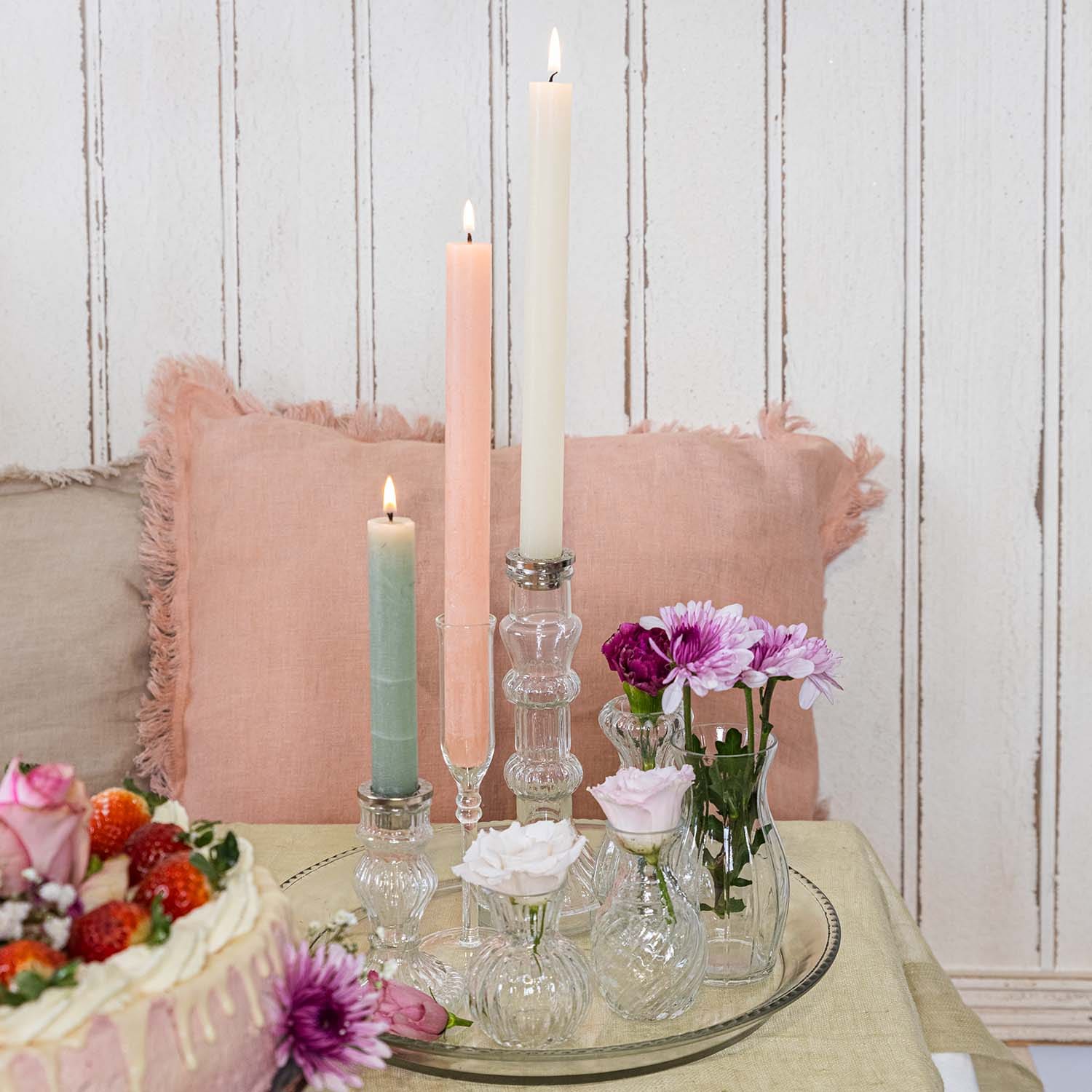 Sada pastelových svíček a skleněných váziček s květinami na skleněném podnosu na stole