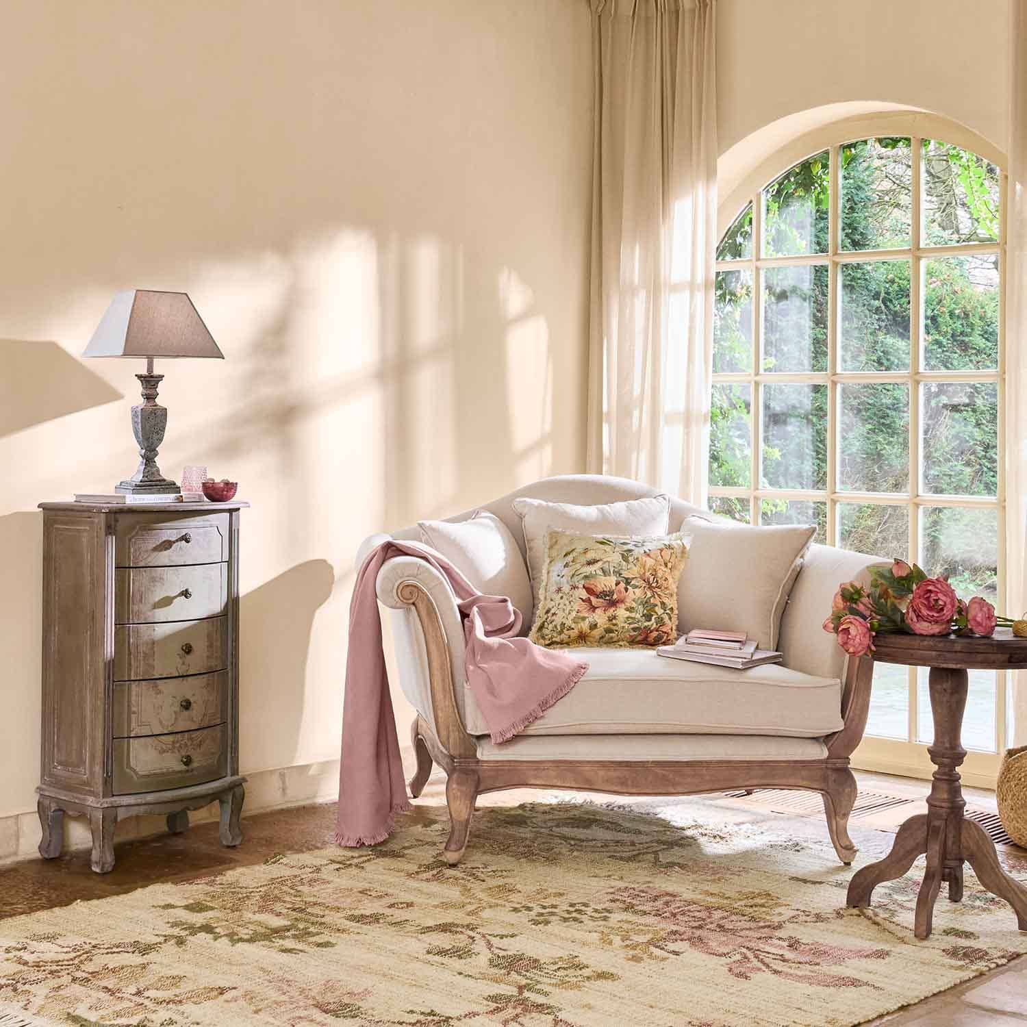 Krémová pohovka s dřevěným rámem, květinový polštář a růžová deka v světlém obývacím pokoji s velkým oknem