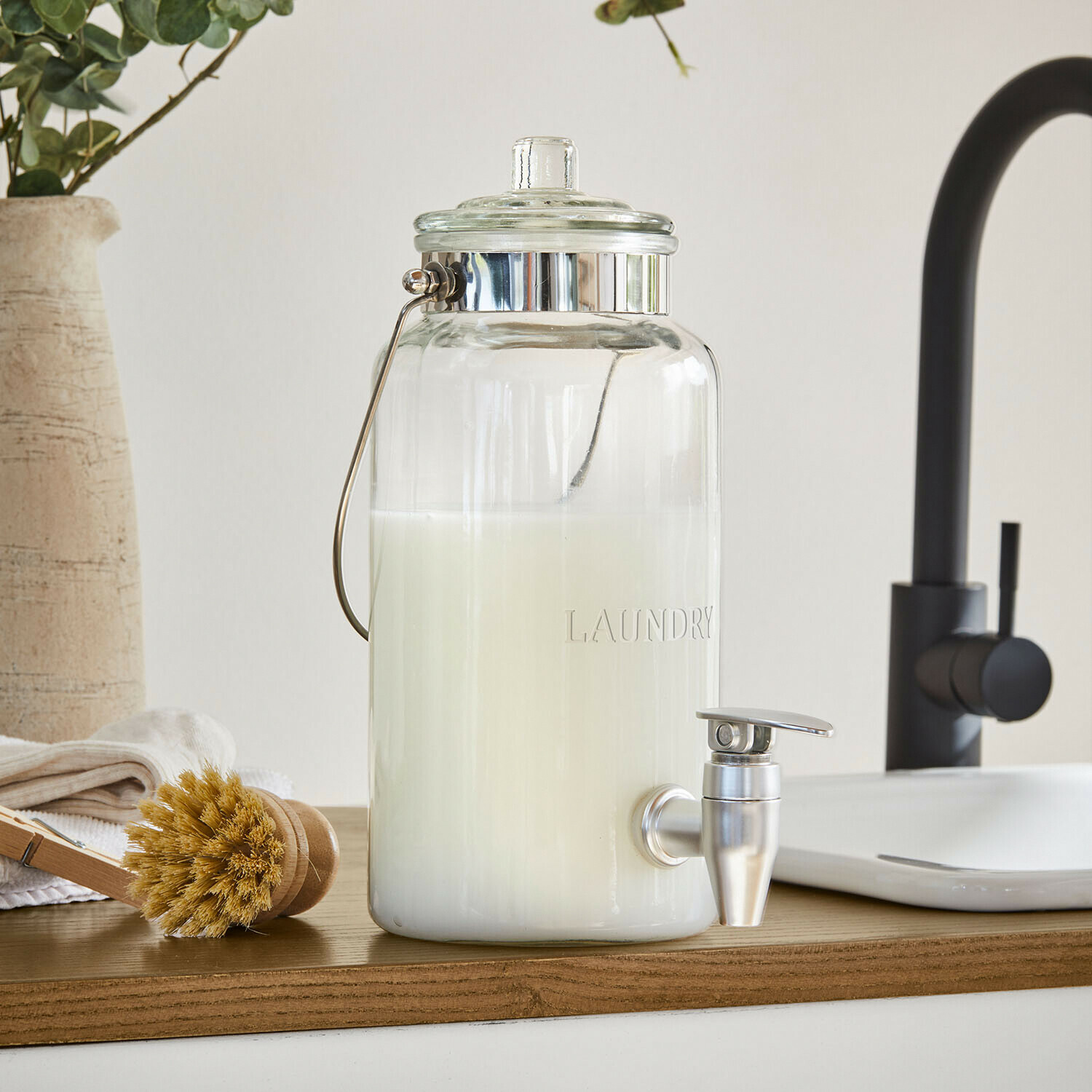 Skleněný dávkovač na prací prostředek s kovovým kohoutkem a držadlem, bílá tekutina uvnitř