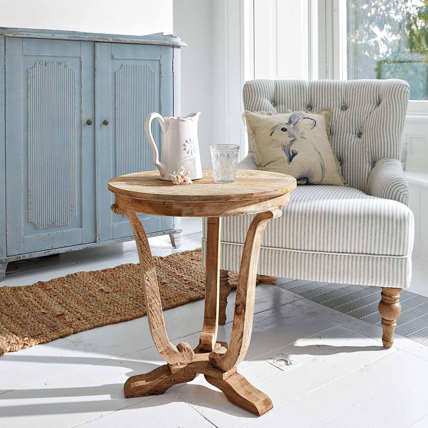 Kulatý stolek z mangového dřeva v přírodní barvě v světlém obývacím pokoji s pruhovaným křeslem a modrou komodou