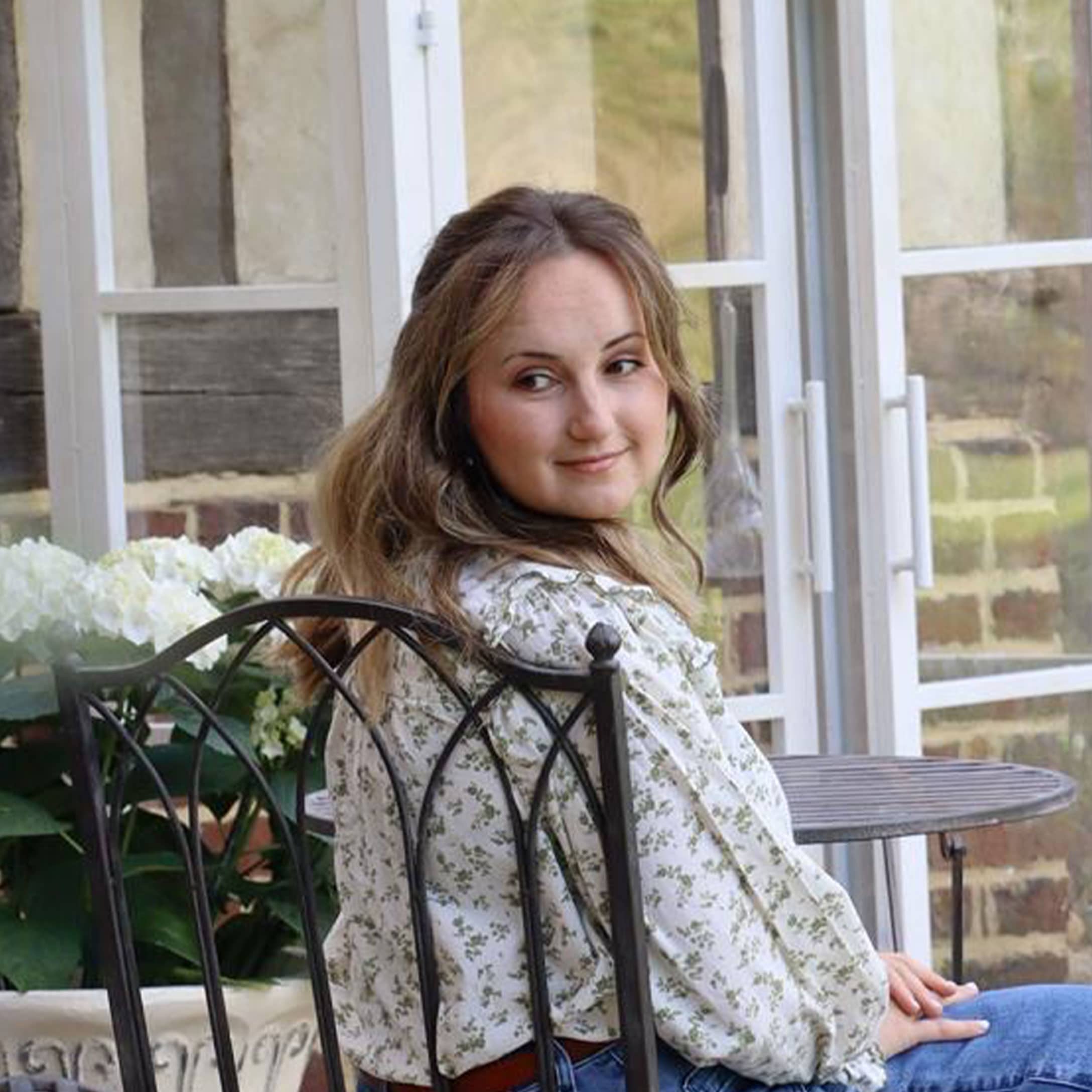 Žena v květované blůze sedí na kovové židli v světlé zahradě s bílými květy