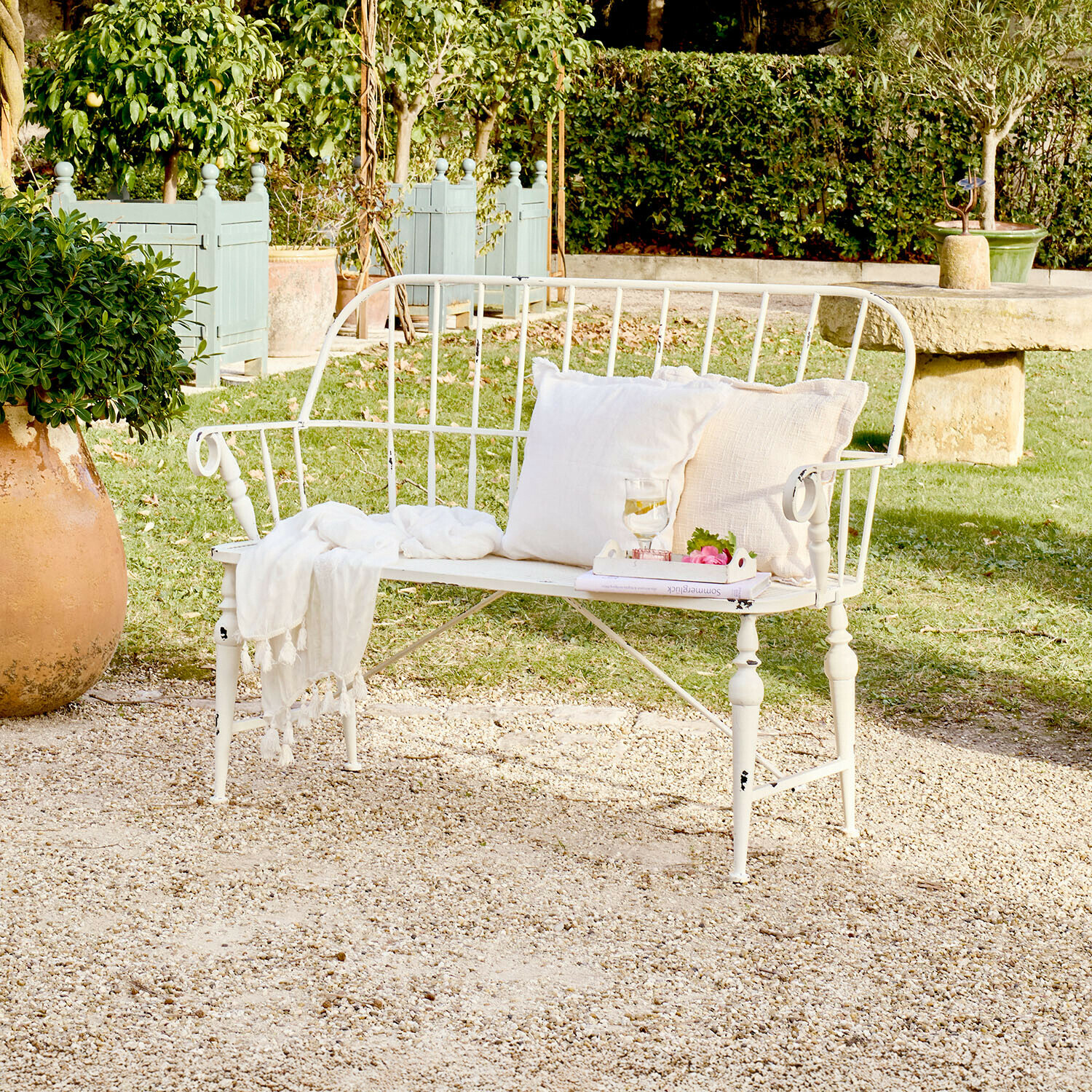 Bílá kovová zahradní lavička s polštáři a dekou na štěrkové terase v zelené zahradě