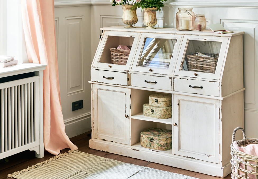 Bílá dřevěná komoda ve stylu shabby chic se skleněnými zásuvkami a dekoracemi v pokoji.