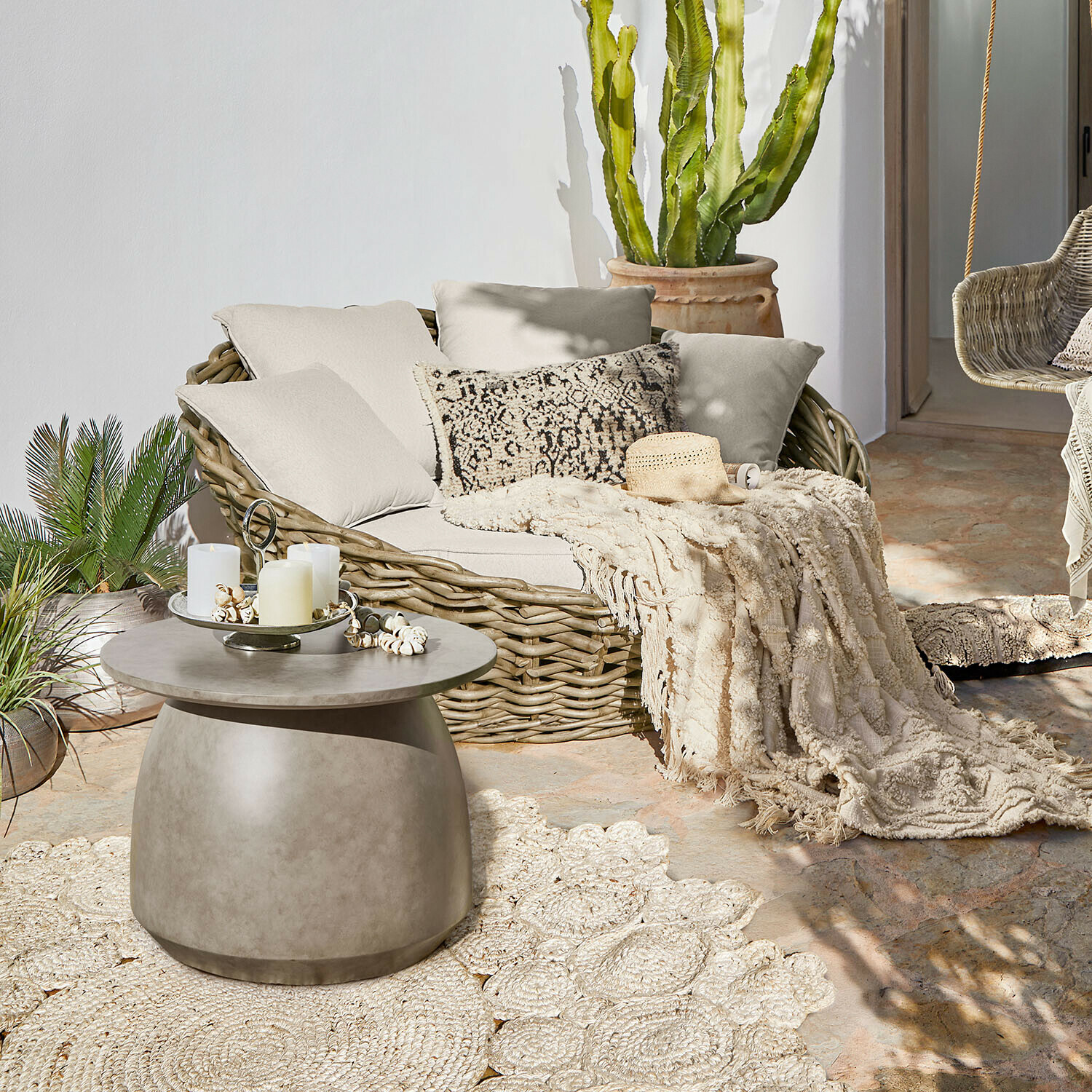Kulatá ratanová pohovka s krémovými polštáři a dekou na terase s betonovým stolkem