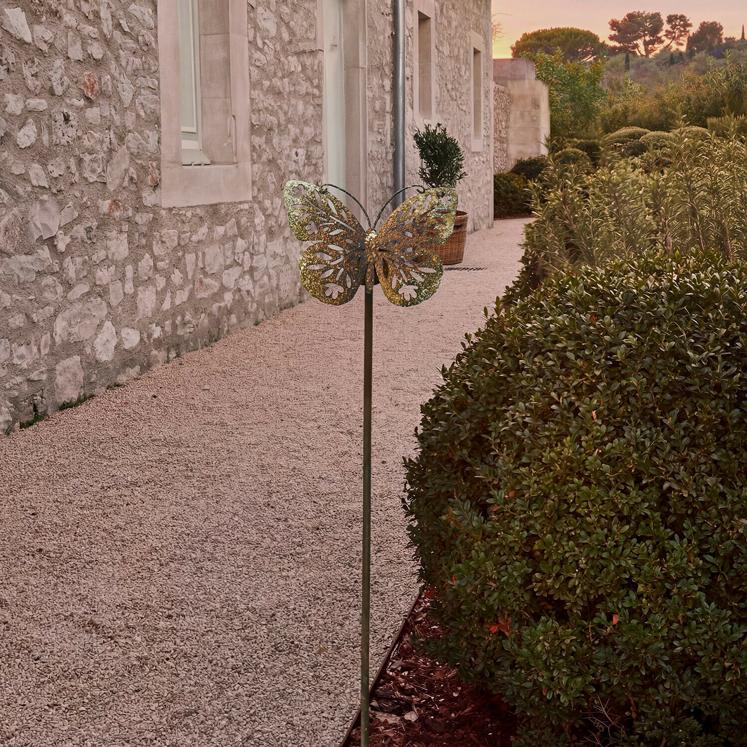 Kovová zahradní dekorace ve tvaru motýla v mosazné barvě před kamennou zdí a zelení