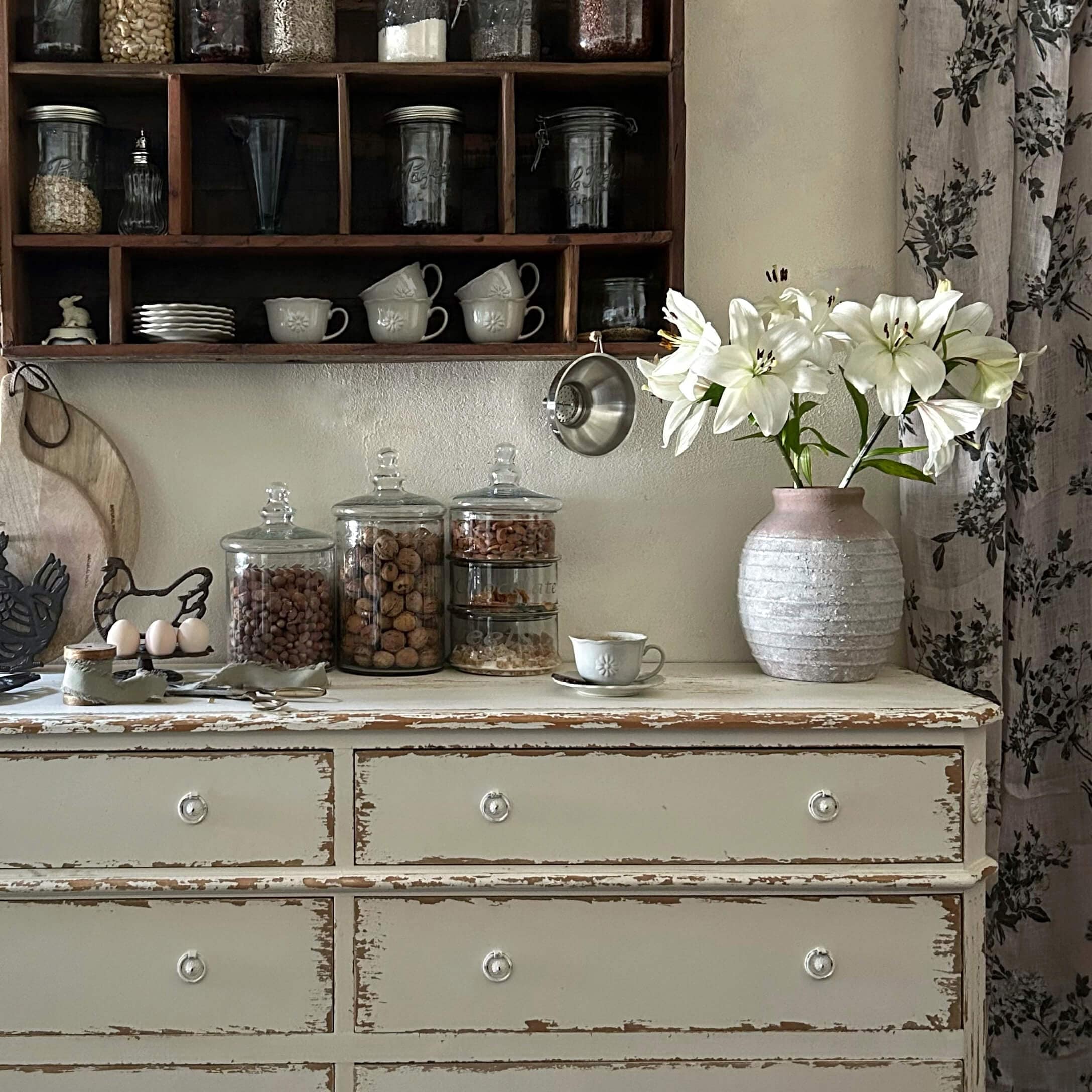 Rustikální bílá komoda s růžovou keramickou vázi a bílými liliemi ve vintage kuchyni