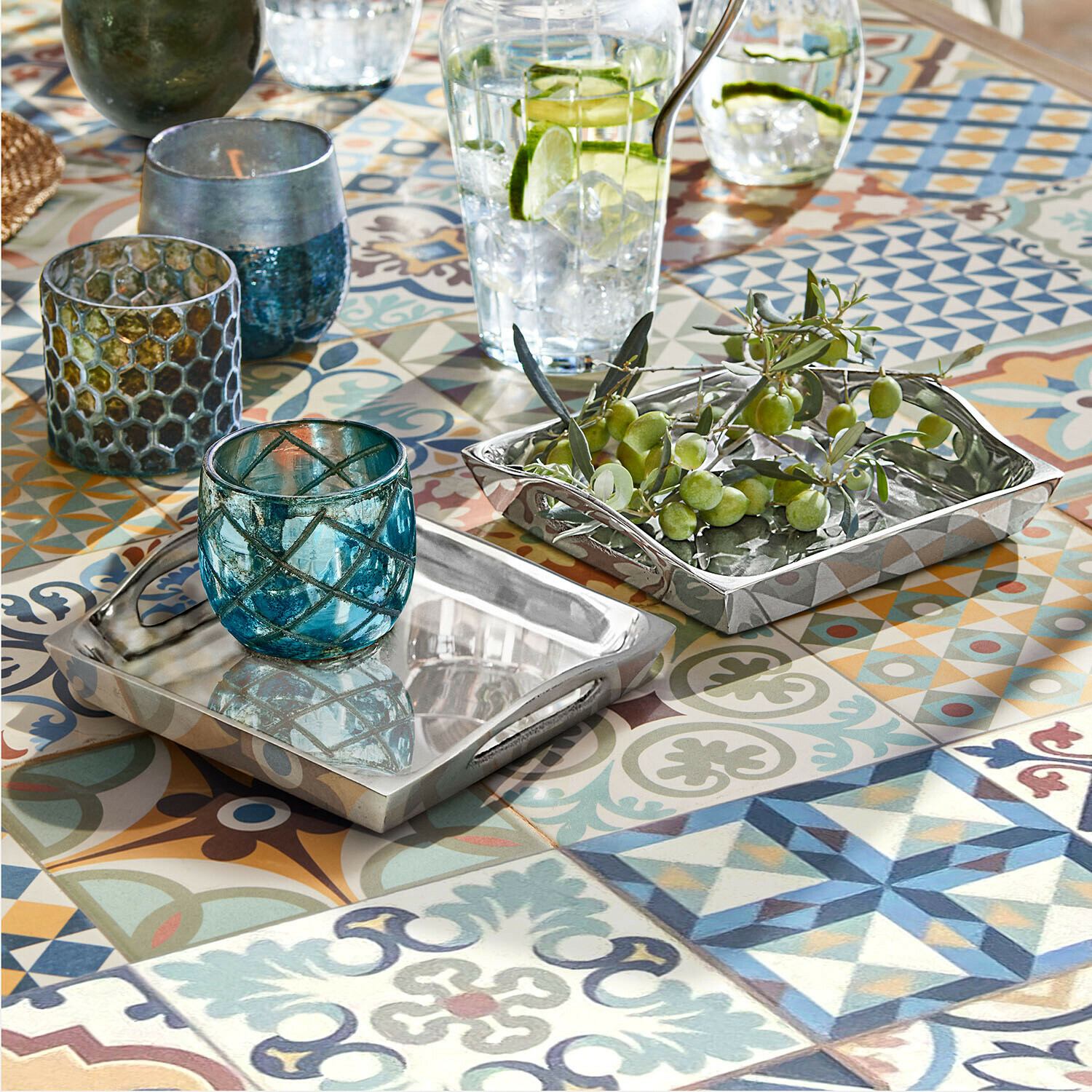 Oboustranné stříbrné kovové podnosy na barevném stole s mozaikou, modré skleněné svícny a zelené olivy