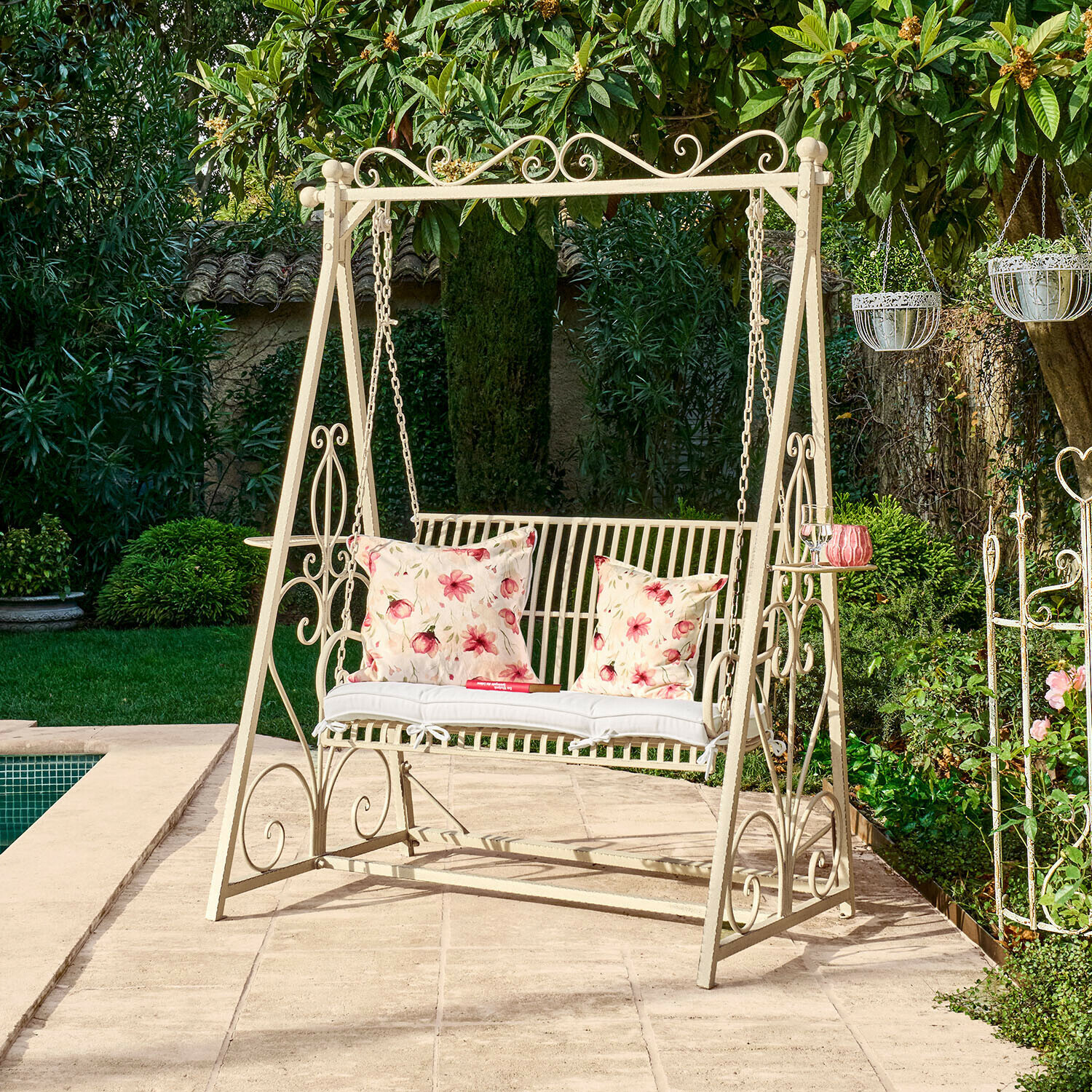 Kovová zahradní houpačka krémové barvy s květinovými polštáři na terase u bazénu