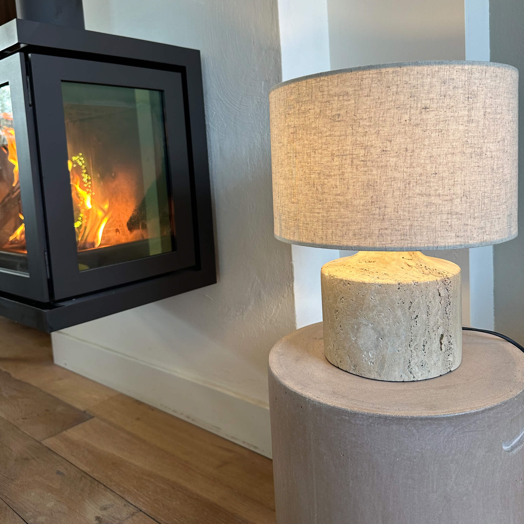 Stolní lampa s béžovým stínidlem a světlým kamenným podstavcem vedle hořícího krbu