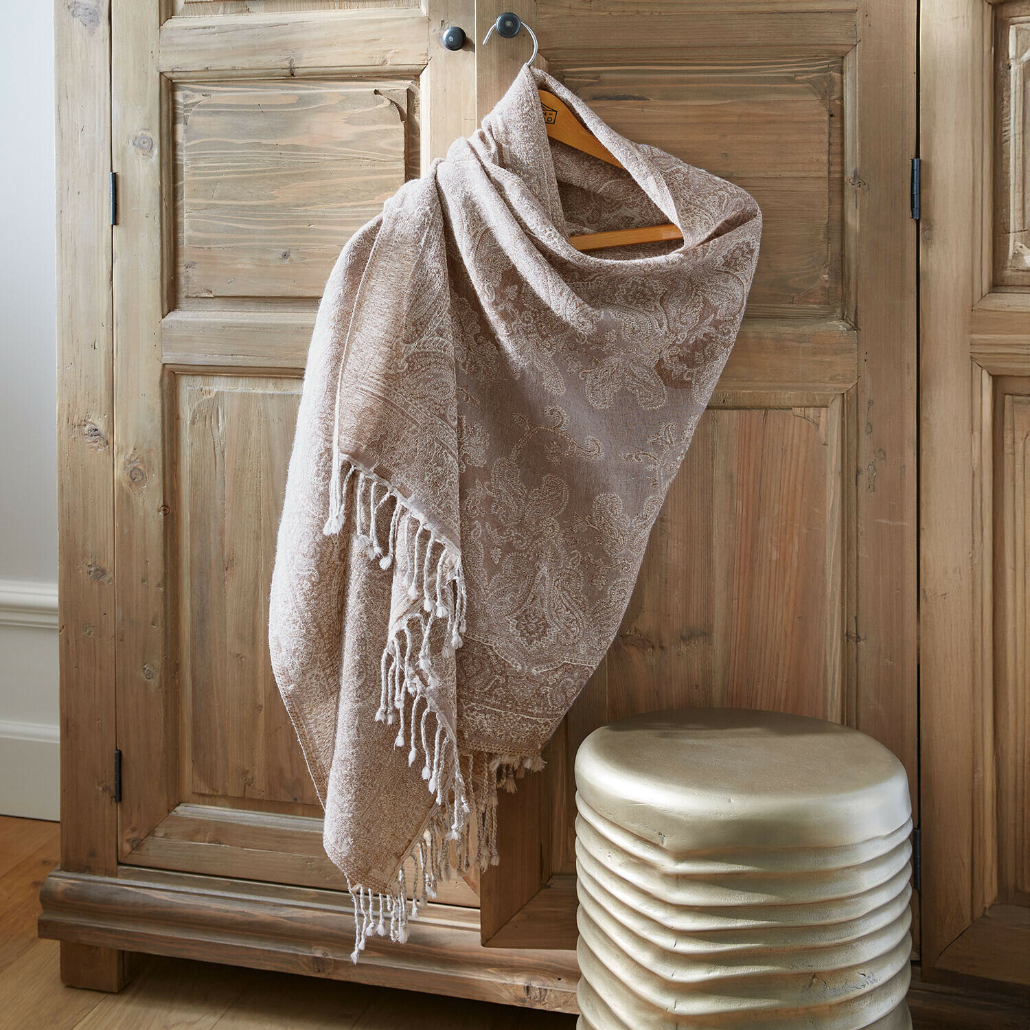 Béžový šál s paisley vzorem a třásněmi zavěšený na dřevěné skříni, měkký materiál, dekorativní doplněk