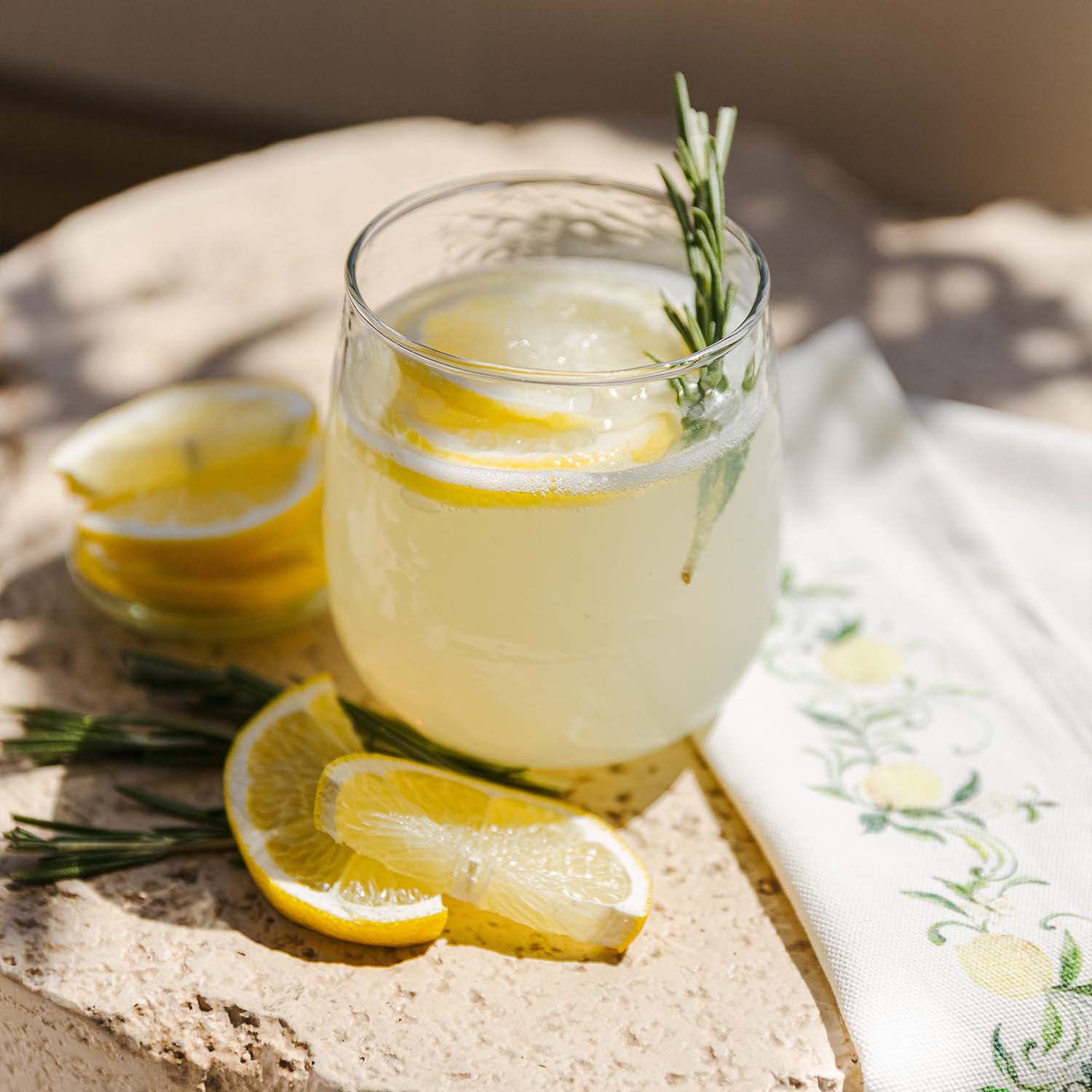 Sklenice citronády s plátky citronu a větvičkou rozmarýnu na kamenném povrchu s ubrouskem