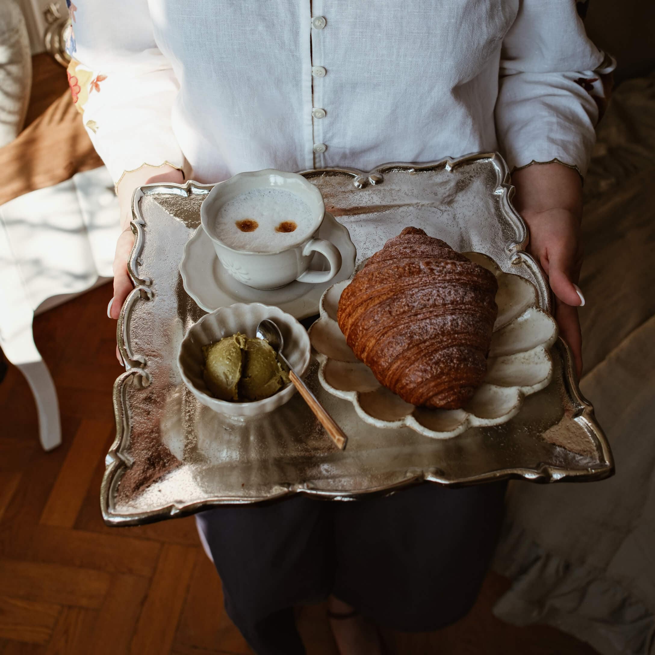 Kovový podnos s croissantem, šálkem kávy a zeleným máslem na bílé porcelánové sadě
