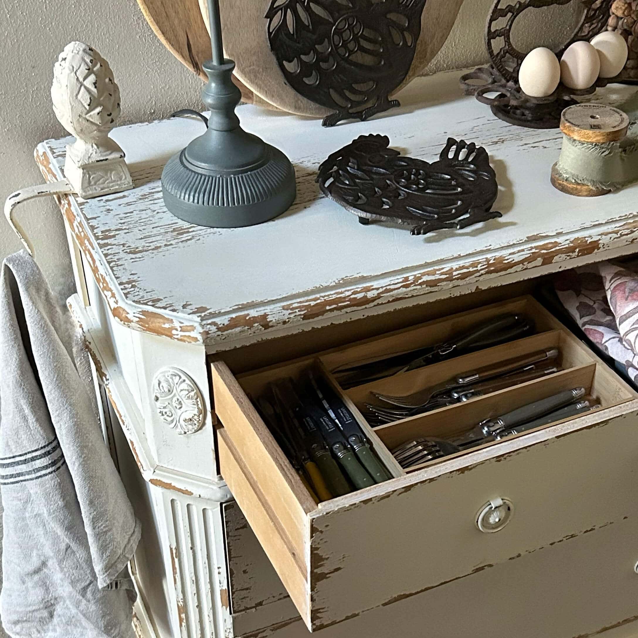 Bílá dřevěná komoda ve stylu vintage s otevřenou zásuvkou a příbory v organizéru, rustikální kuchyně