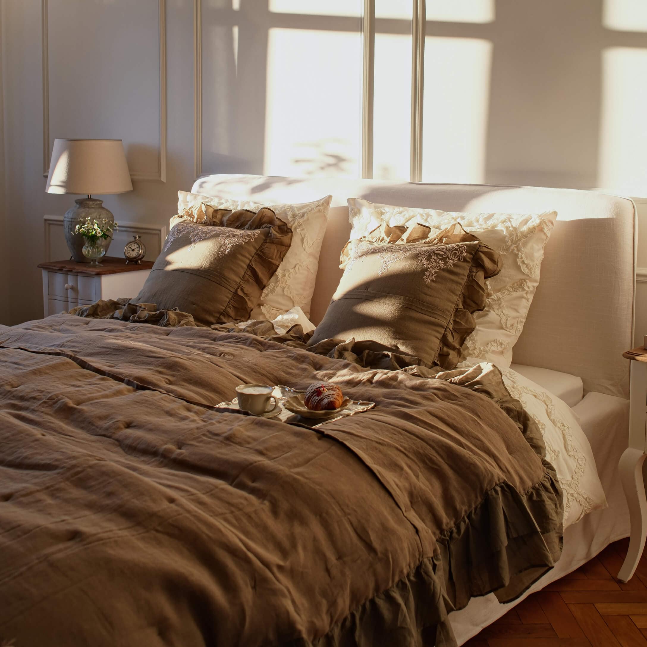 Pohodlná ložnice s béžovým lněným povlečením a hnědými polštáři s volánky ve slunečním světle