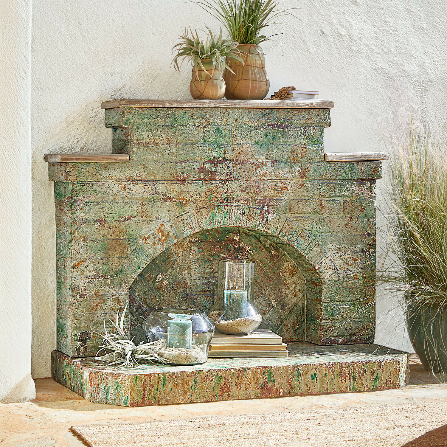 Dekorativní krb z patinovaného kovu v zeleno-rezavých odstínech se svíčkami a rostlinami
