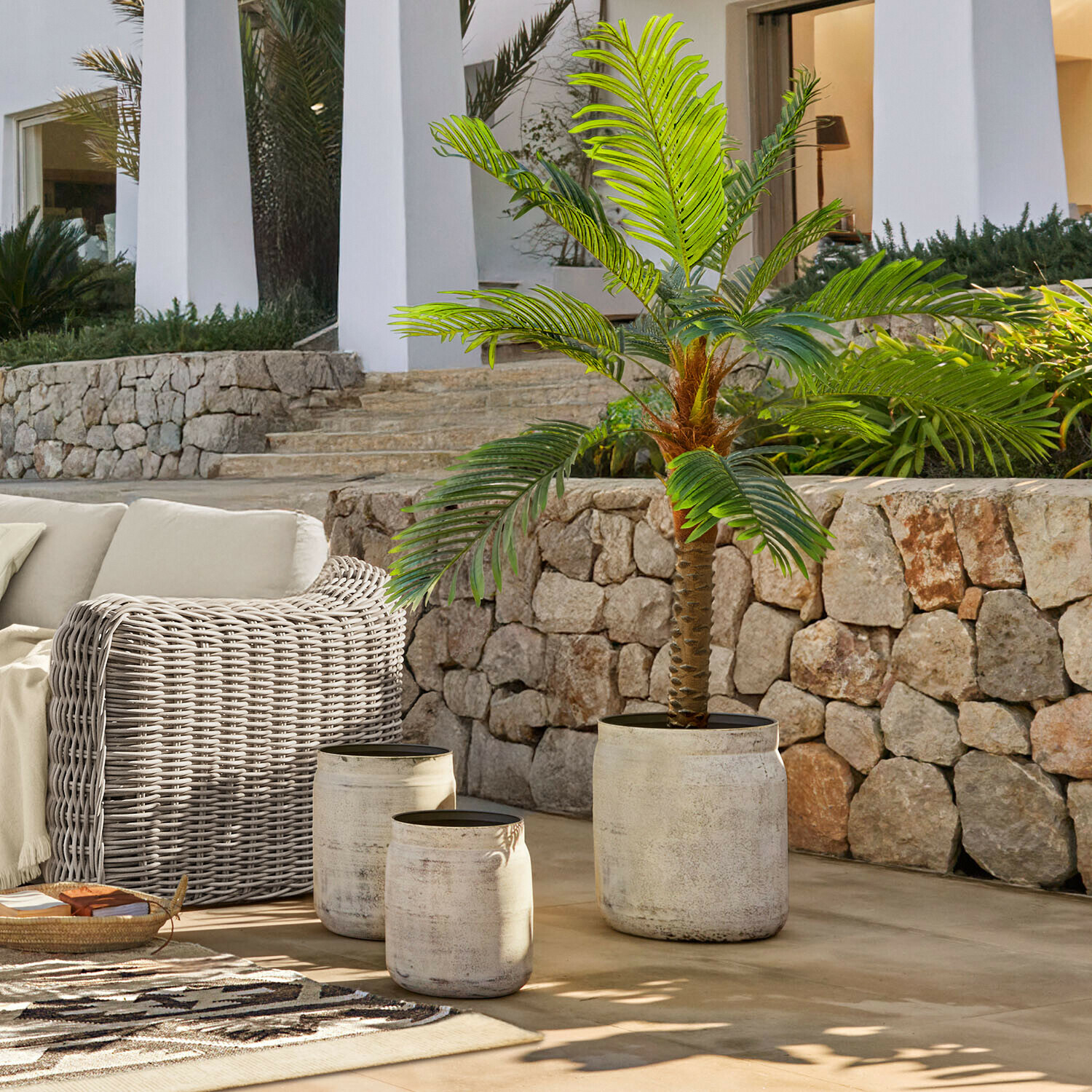 Sada tří světlých betonových květináčů na terase se zelenou umělou palmou a ratanovým nábytkem