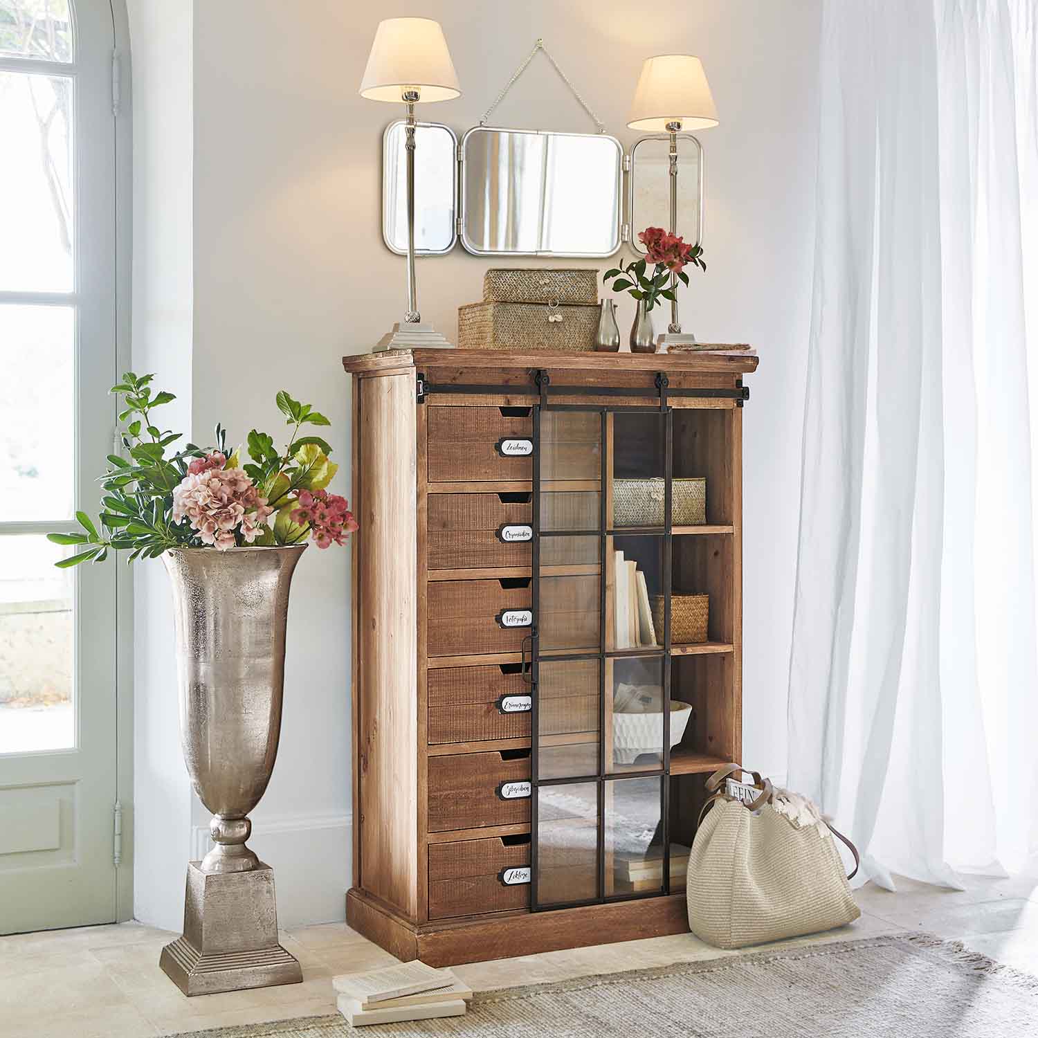 Dřevěná komoda se zásuvkami a skleněnými dveřmi v světlém interiéru s dekoracemi a lampami