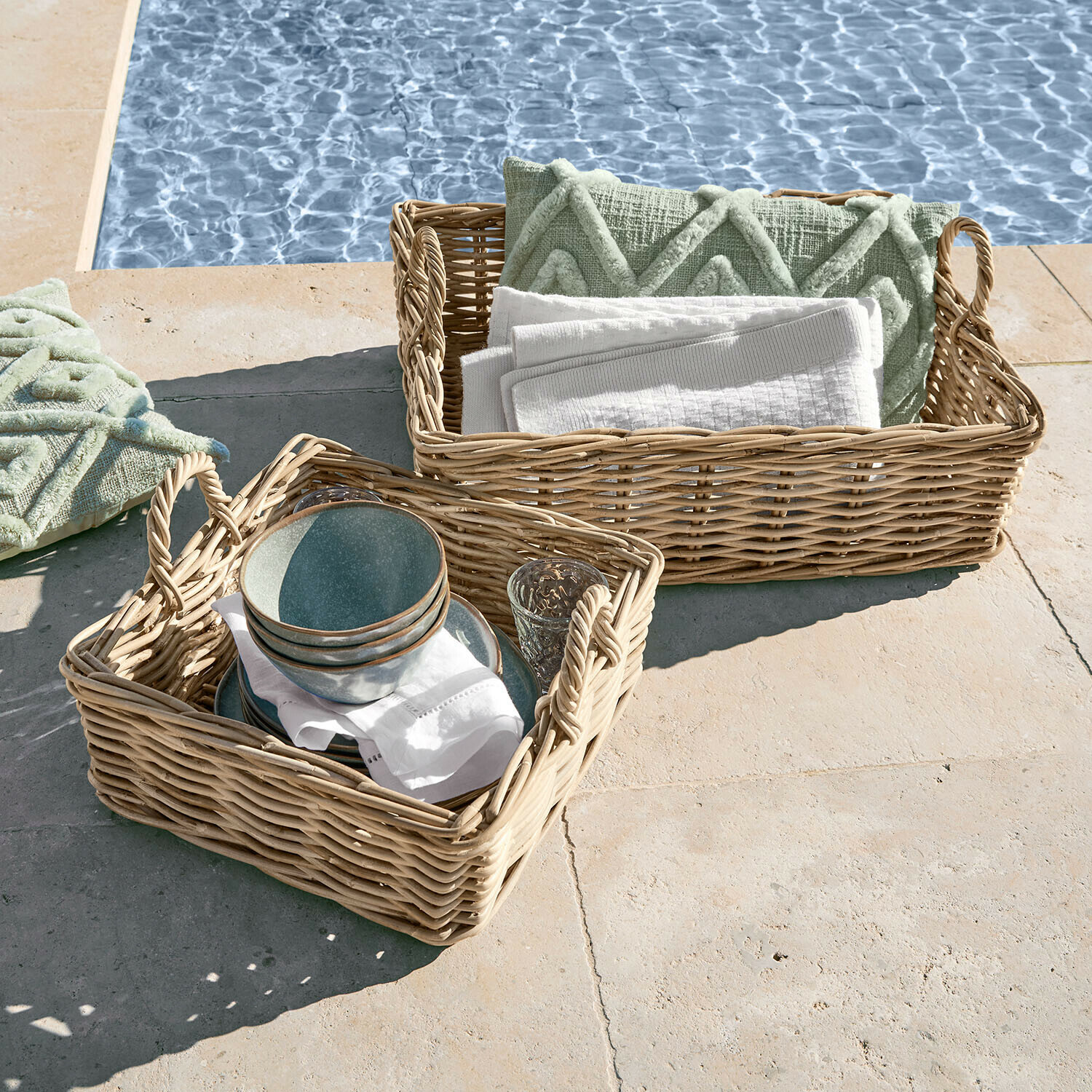 Dva proutěné koše s úchyty, naplněné zelenými polštáři a bílými ručníky u bazénu