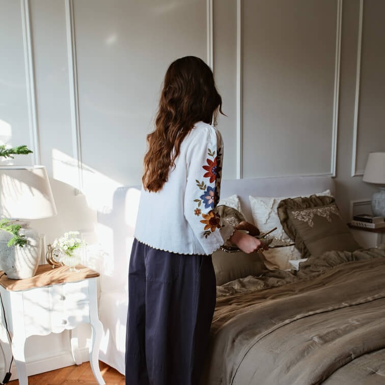 Žena v bílé blůze s barevnou výšivkou na rukávech v ložnici s hnědým povlečením a lampou na nočním stolku