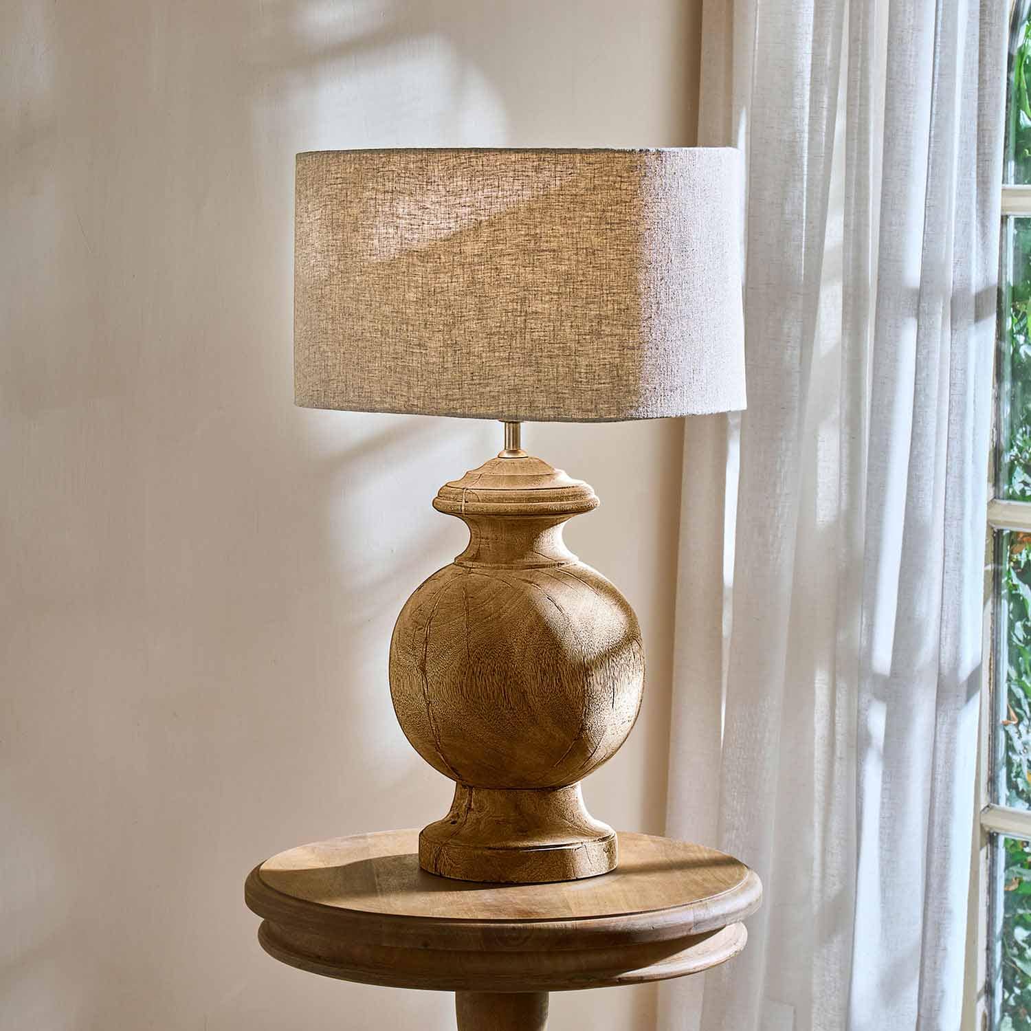 Stolní lampa s dřevěnou základnou a béžovým stínidlem na kulatém stolku u okna