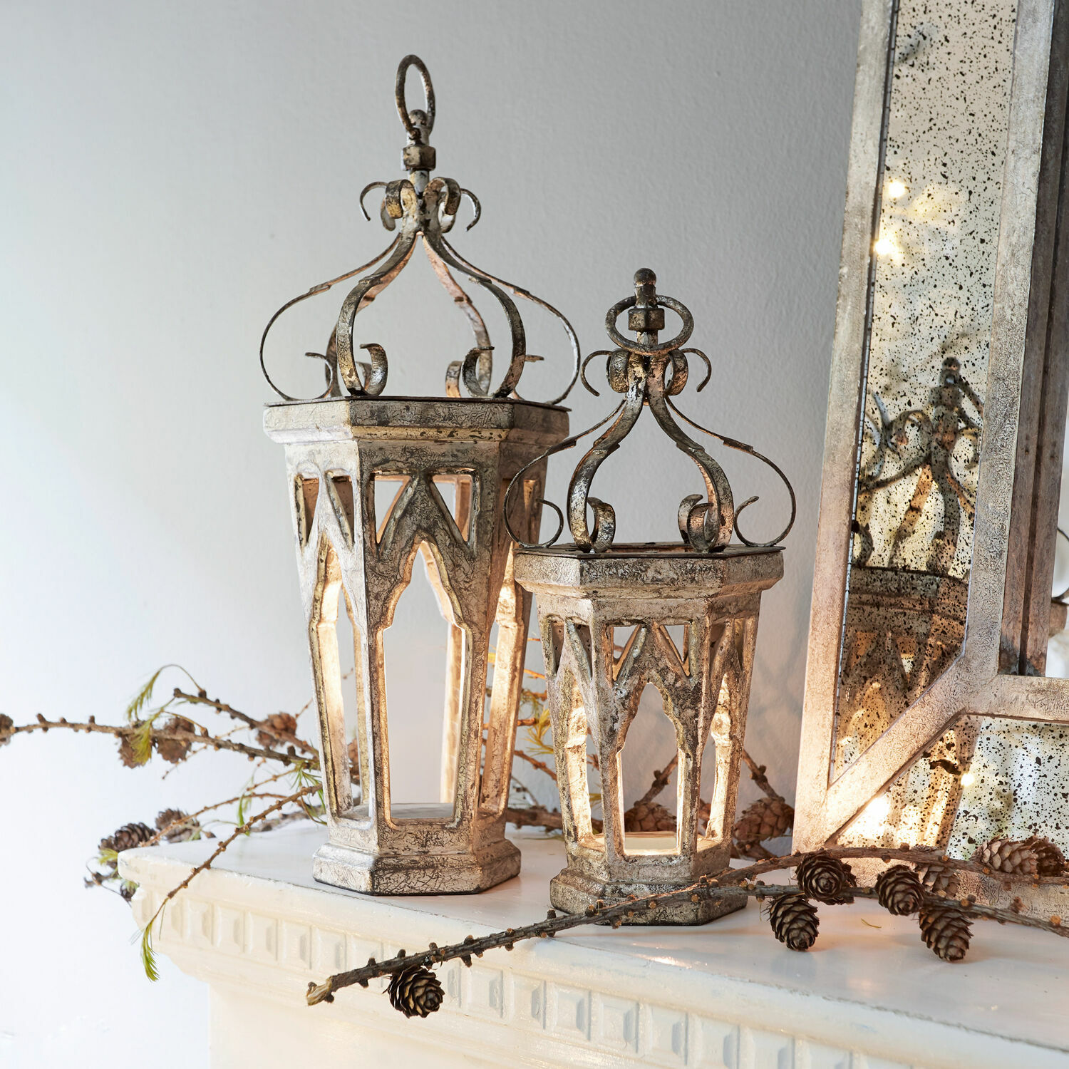 Dekorativní kovové lucerny ve vintage stylu v barvě starostříbra na krbové římse s šiškami