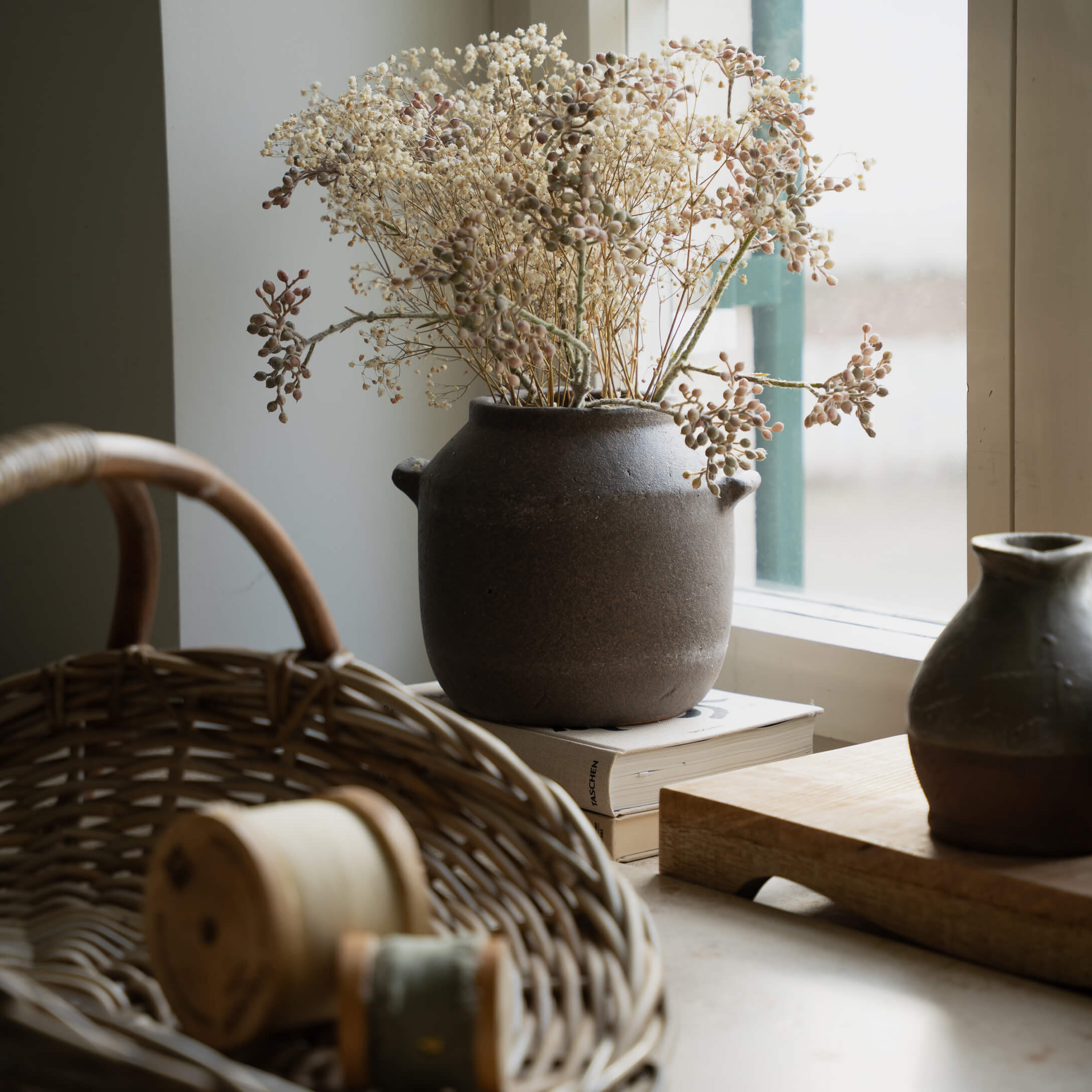 Hnědá keramická váza se sušenými květinami na parapetu vedle proutěného koše a knih
