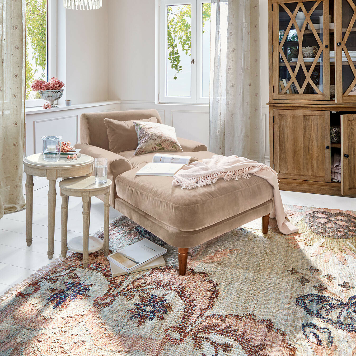 Béžové sametové ležení s polštáři a růžovou dekou na vzorovaném koberci v světlém obývacím pokoji