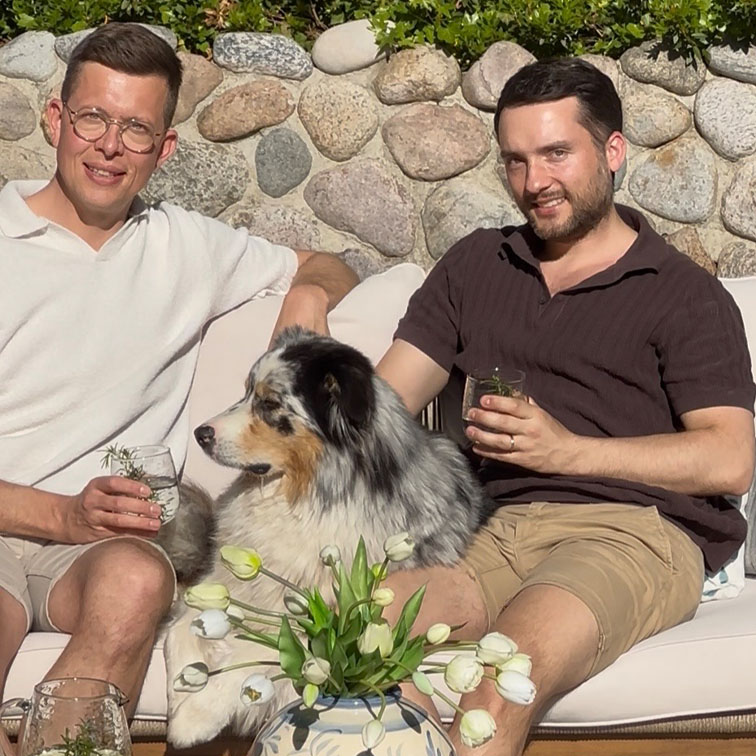 Dva muži sedí venku na pohovce s australským ovčákem a bílými tulipány v květináči.