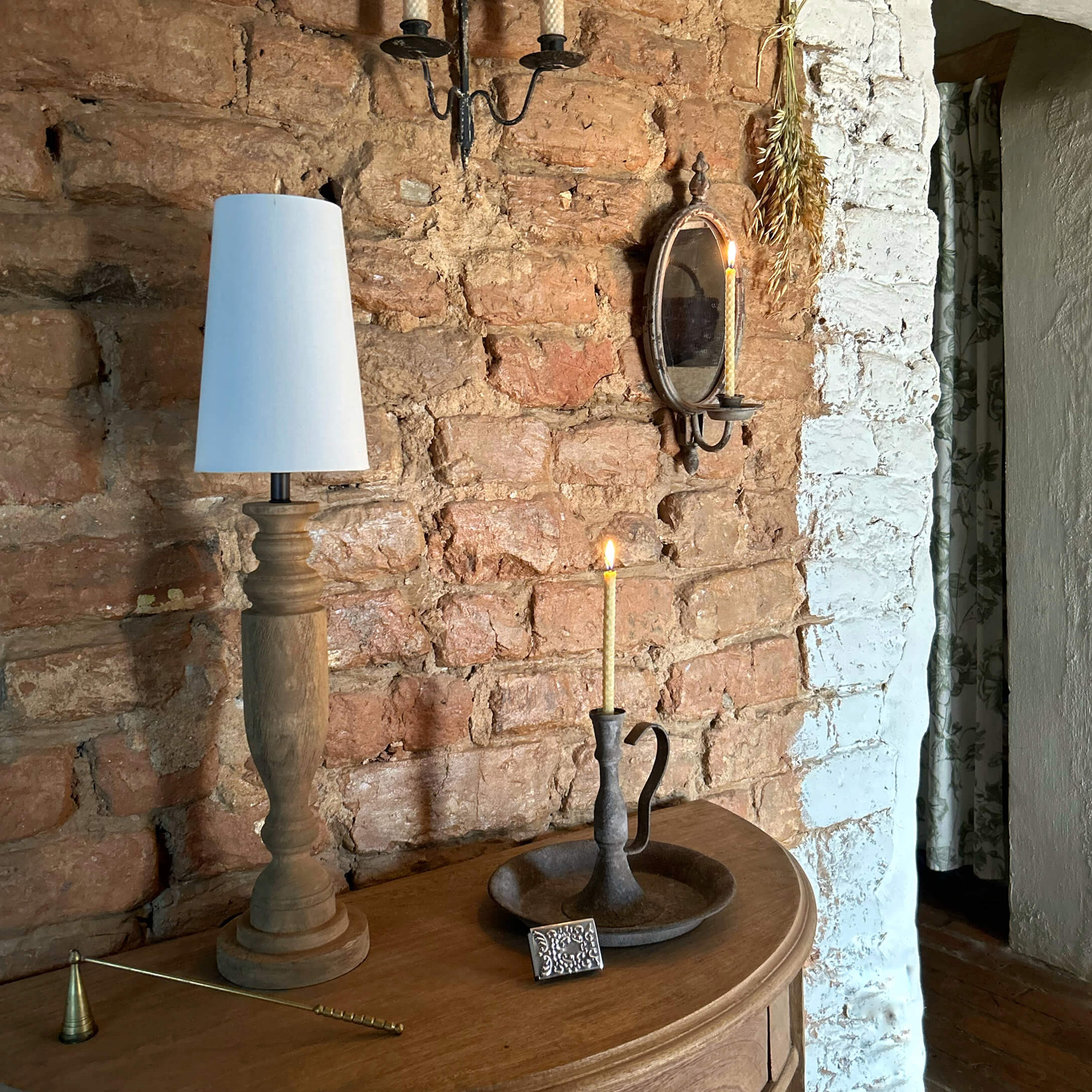 Dřevěná lampa s bílým stínidlem a kovový svícen se zapálenou svíčkou na dřevěném stole u cihlové zdi