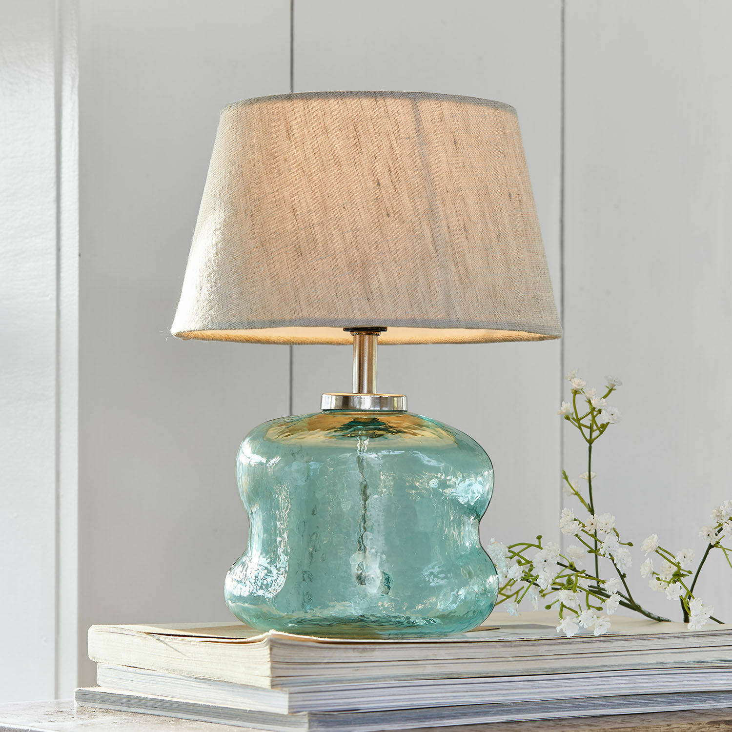 Stolní lampa Rufino Stolní lampa z modrého skla s béžovým stínidlem na hromadě knih, interiérová dekorace