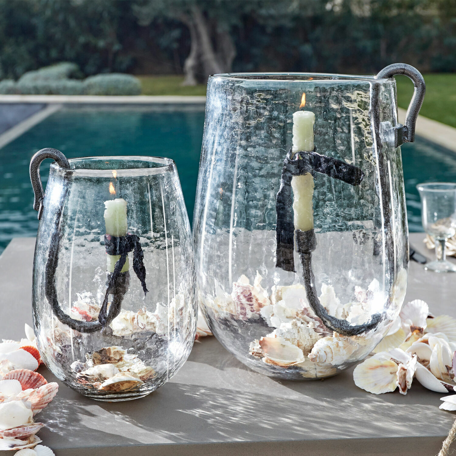 Skleněné lucerny s kovovým držadlem a bílými svíčkami, naplněné mušlemi, venkovní dekorace