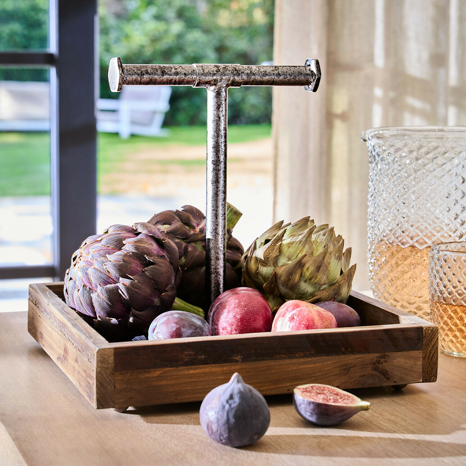 Dřevěný podnos s kovovou rukojetí plný artyčoků a švestek na světlém stole v interiéru