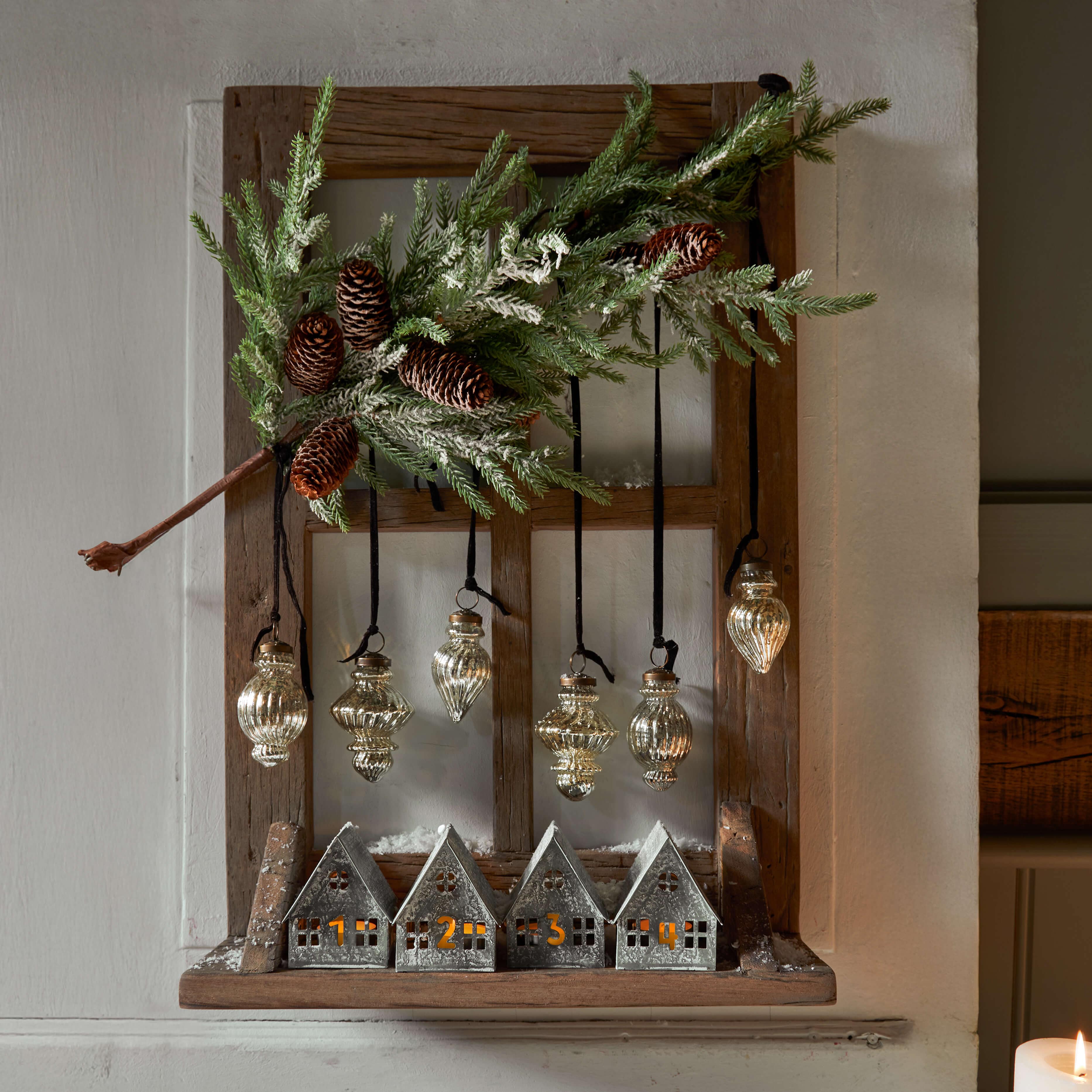 Dřevěný rám s kovovými domečky a závěsnými skleněnými ozdobami, vánoční dekorace s šiškami a zelenými větvičkami