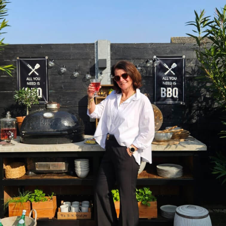 Žena v bílé košili a černých kalhotách u venkovního grilu s nápisem BBQ na černé zdi