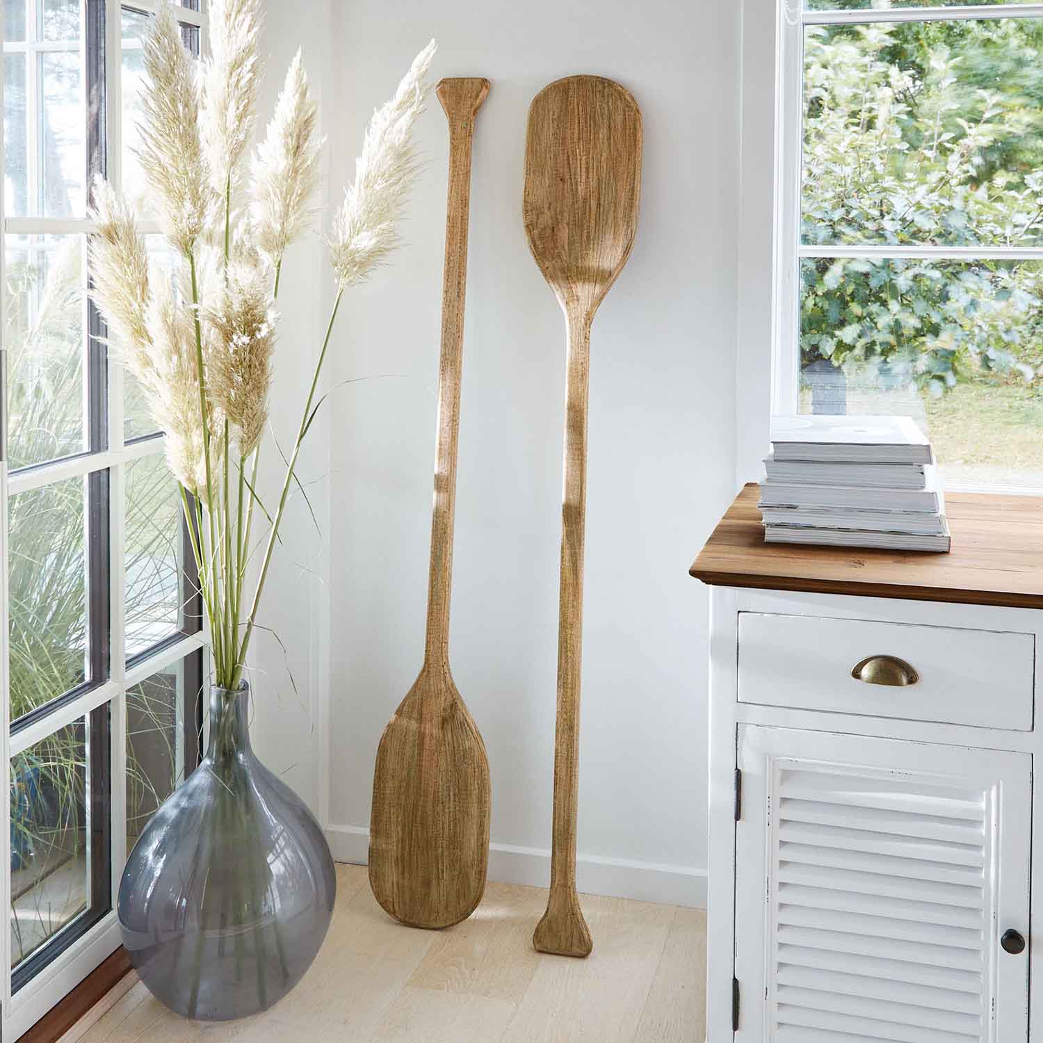 Dvě dekorativní dřevěná pádla v přírodní barvě opřená o bílou zeď v světlém interiéru