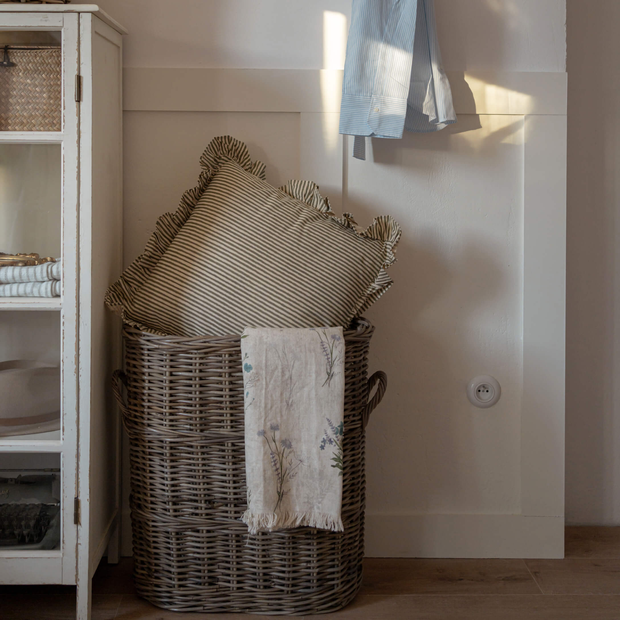 Hnědý proutěný koš s proužkovaným polštářem a lněnou dekou s květinovým vzorem v útulném interiéru