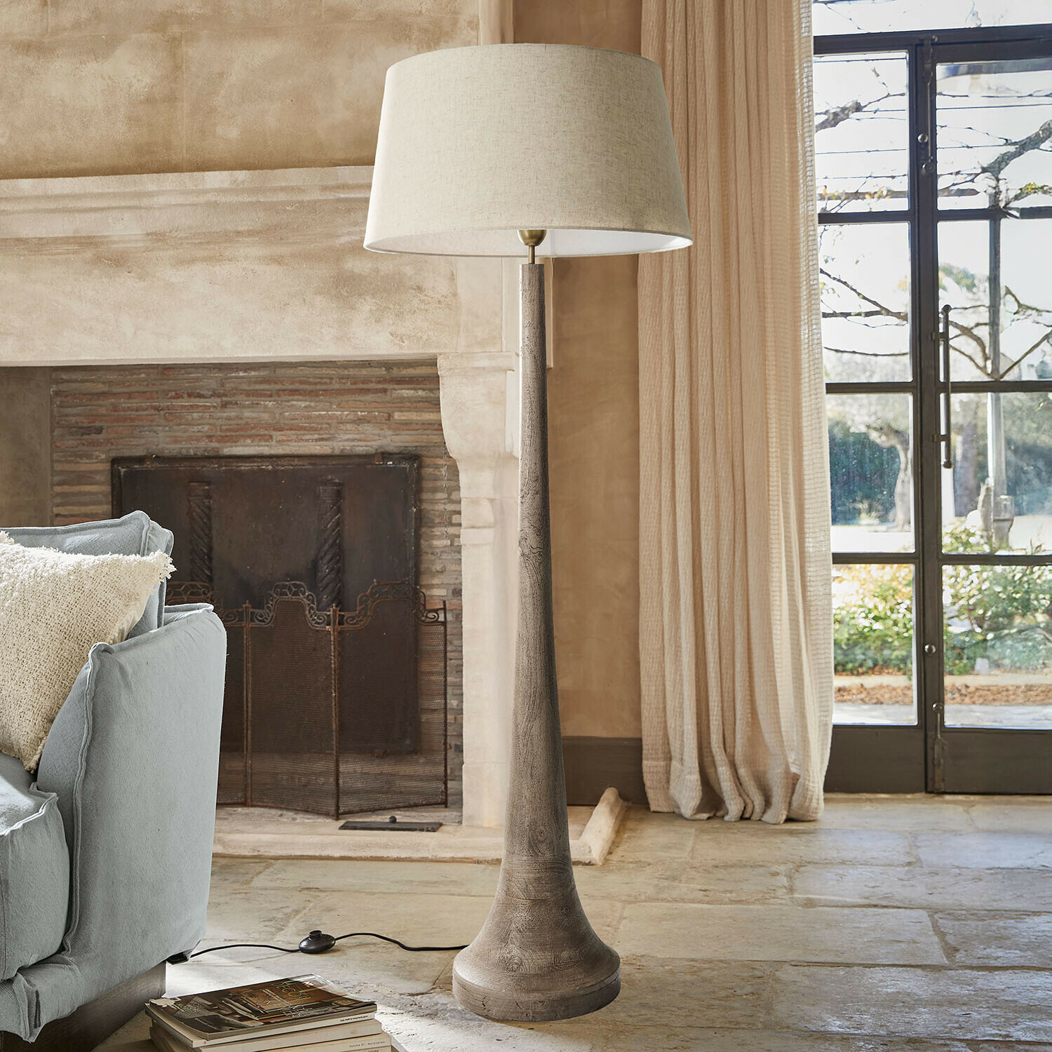 Stojací lampa s dřevěným podstavcem a béžovým stínidlem v útulném obývacím pokoji s krbem