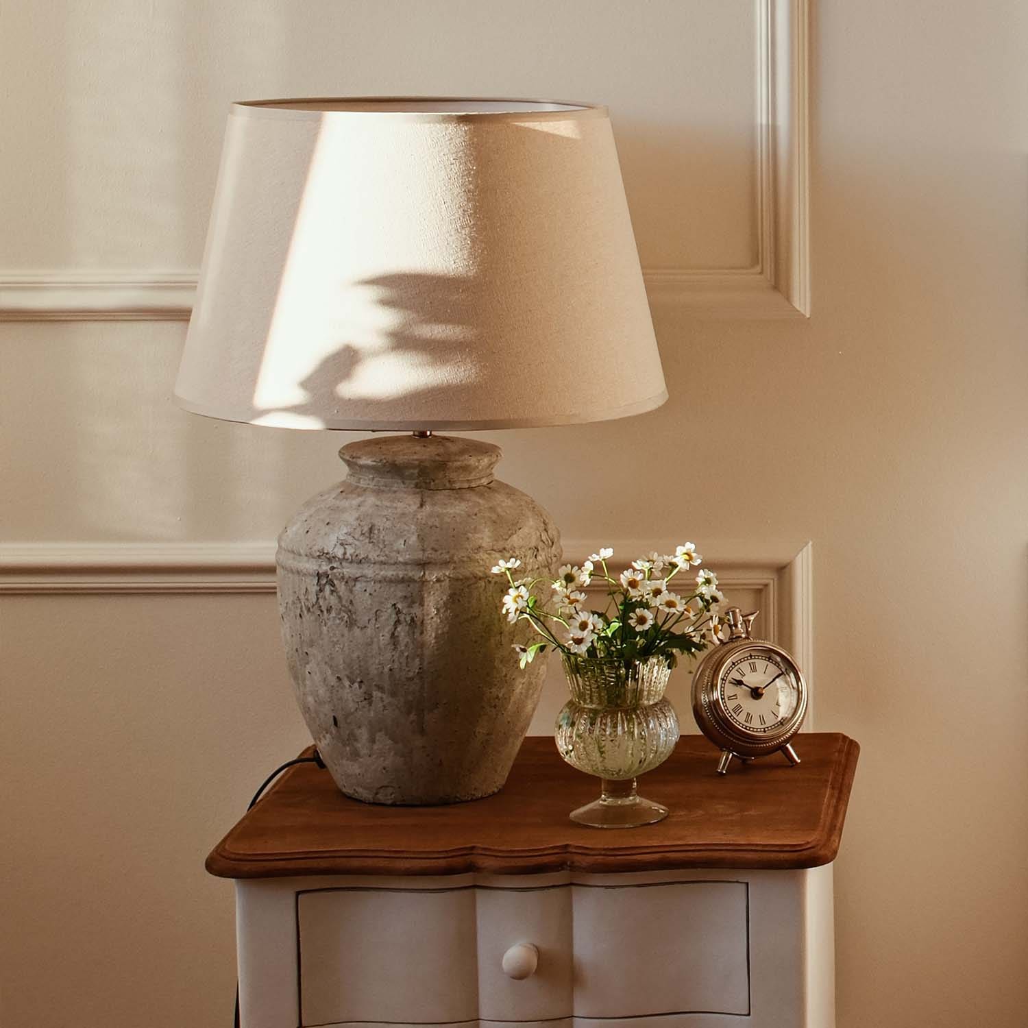 Stolní lampa s béžovým stínidlem a keramickou vintage základnou na dřevěném stolku s květinami a hodinami
