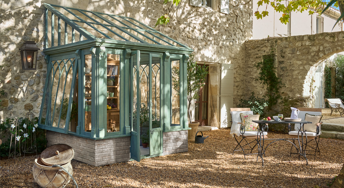 Zelená skleník s kovovým rámem na kamenném podstavci na slunném dvoře s kovovým zahradním nábytkem